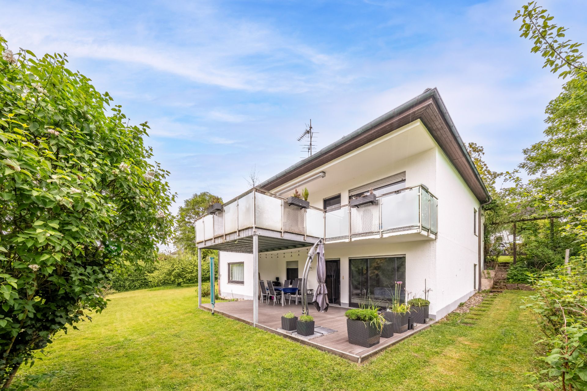 Modernes zweistöckiges Wohnhaus mit Balkon, Terrasse und Garten, umgeben von Bäumen.