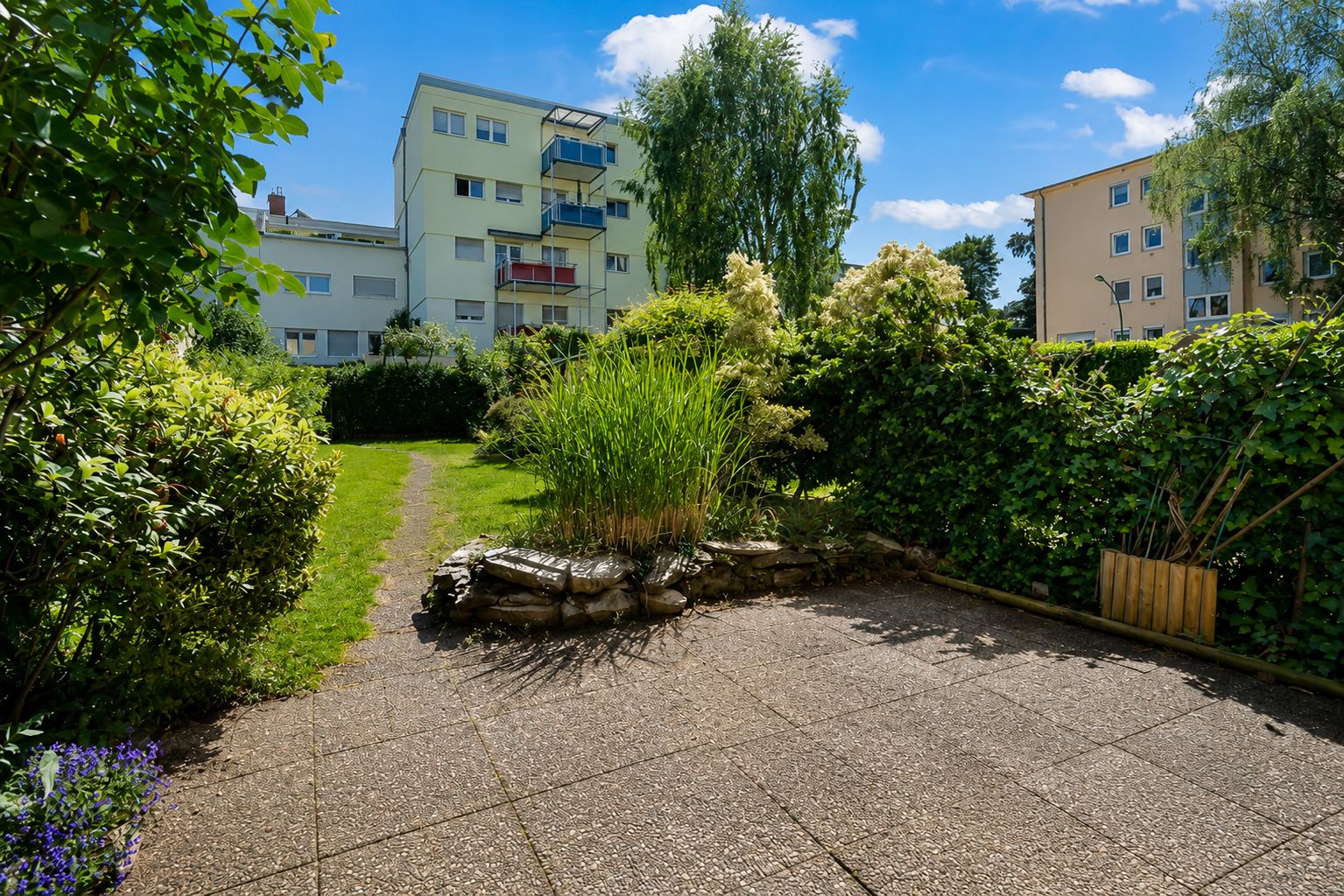 Gepflasterte Terrasse mit Blick auf Garten und mehrstöckige Wohnhäuser im Hintergrund.