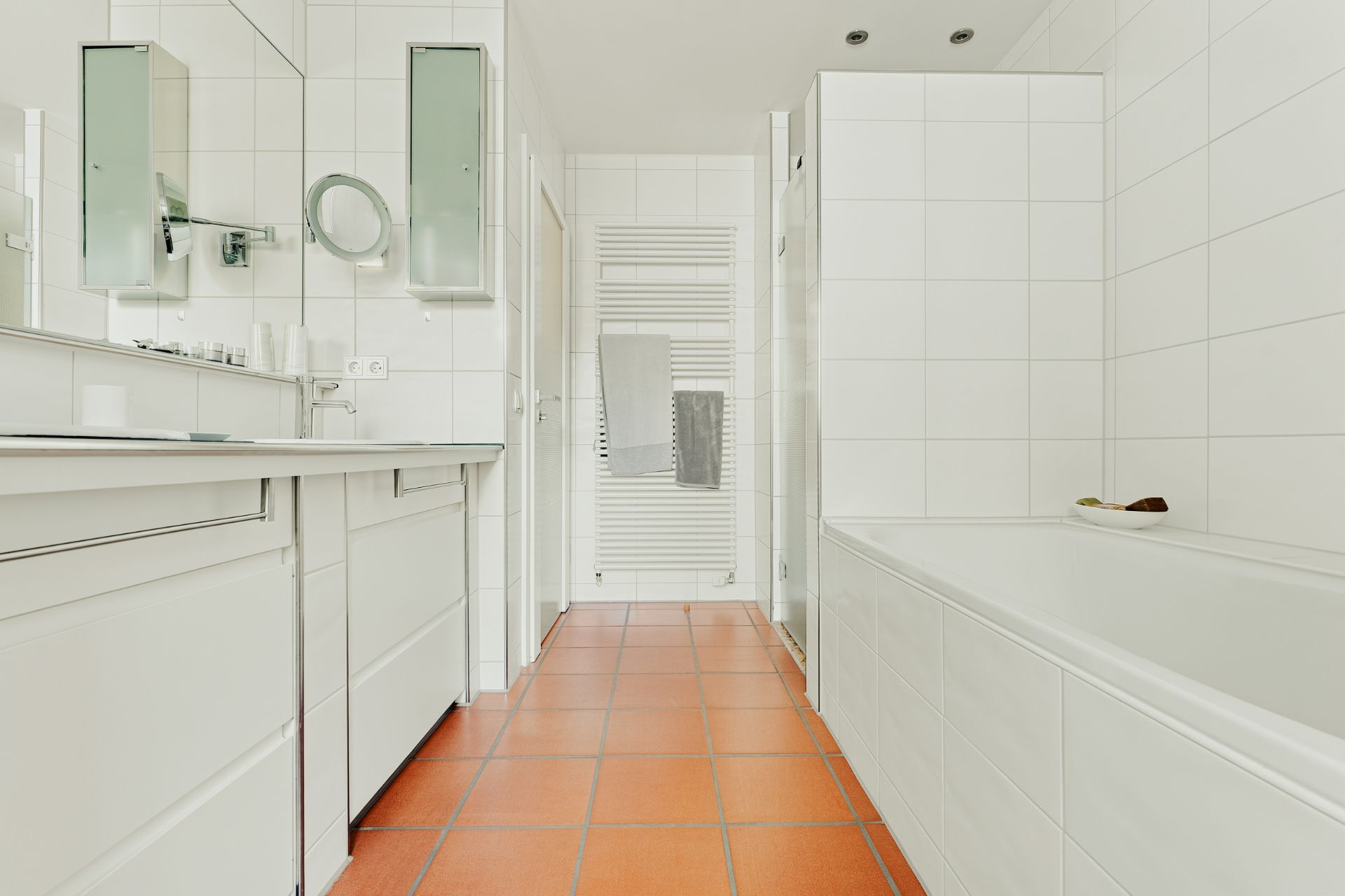 Helles Badezimmer mit Badewanne, zwei Spiegeln, Waschbecken und Handtüchern auf einem Heizkörper.