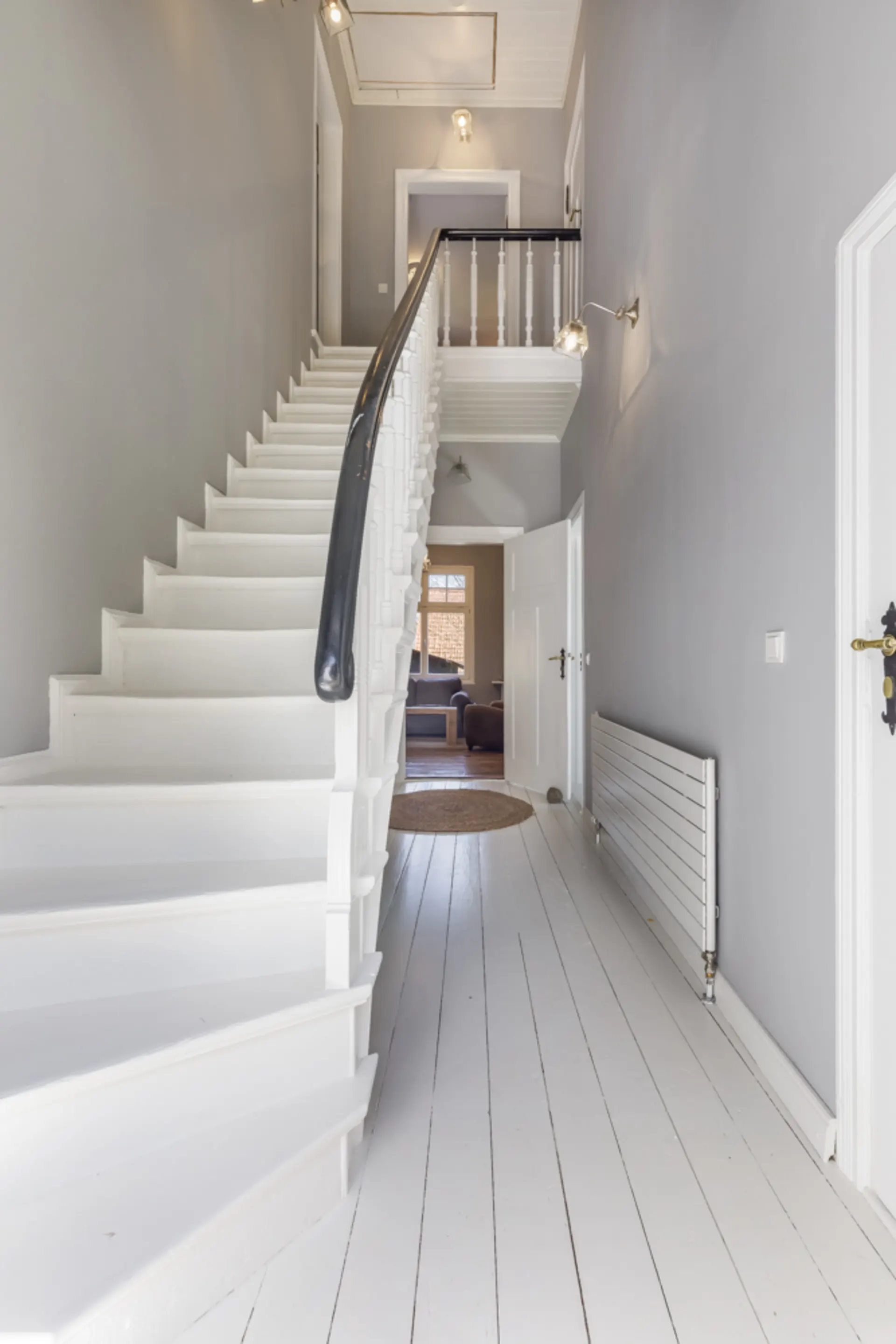 Heller Flur mit weißer Treppe, Holzboden und Blick in ein Wohnzimmer.