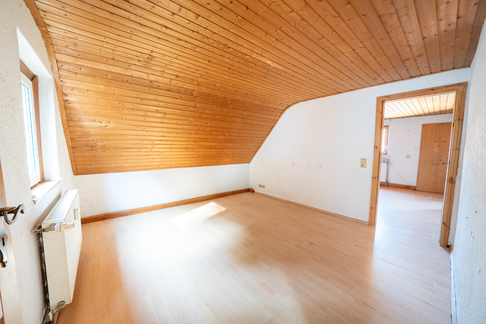 Helles Zimmer mit schräger Holzdecke, hellem Boden und großem Fenster.