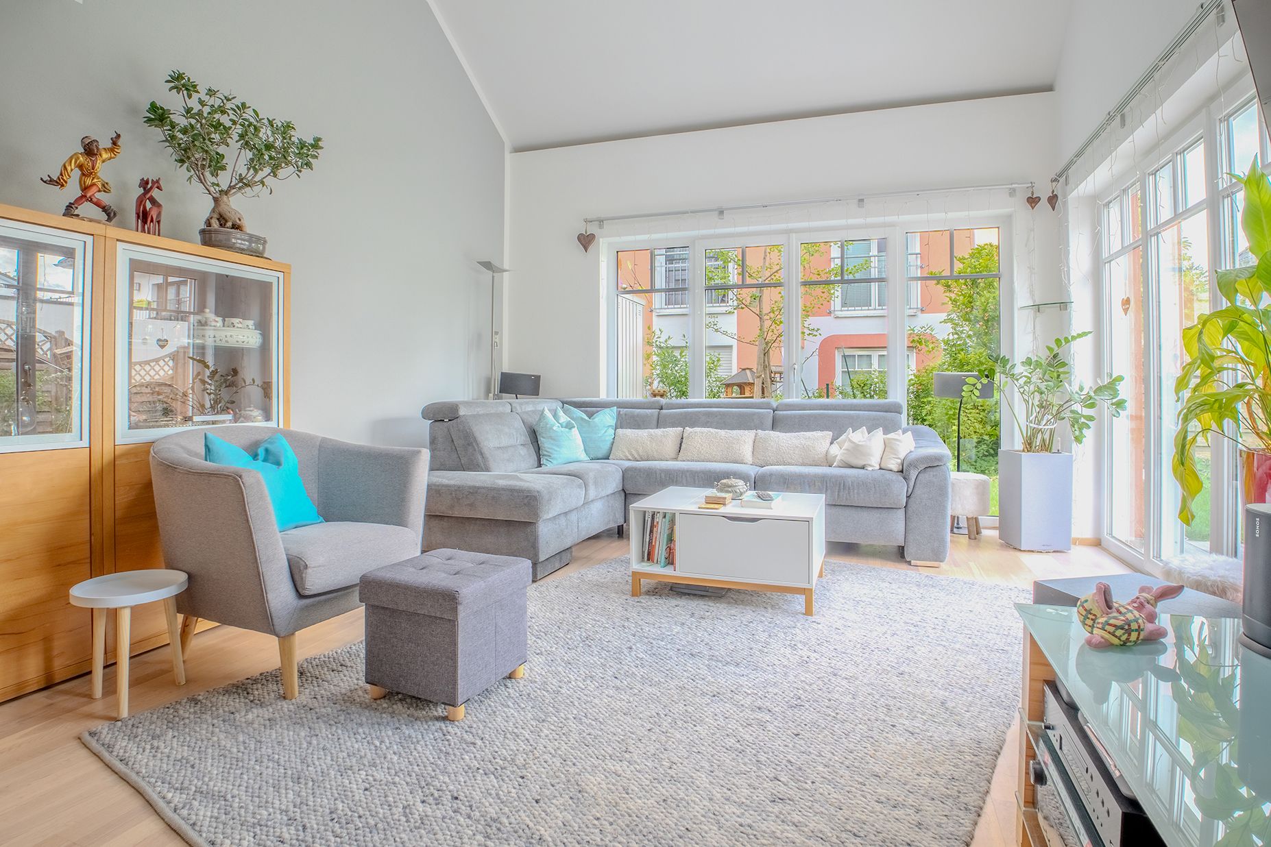 Helles Wohnzimmer mit grauem Sofa, großem Teppich, Pflanzen und großen Fenstern.
