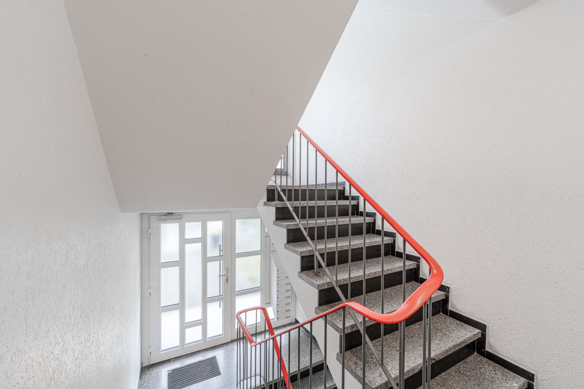 Treppenhaus mit grauen Stufen, rotem Geländer und Glastür am Eingang.