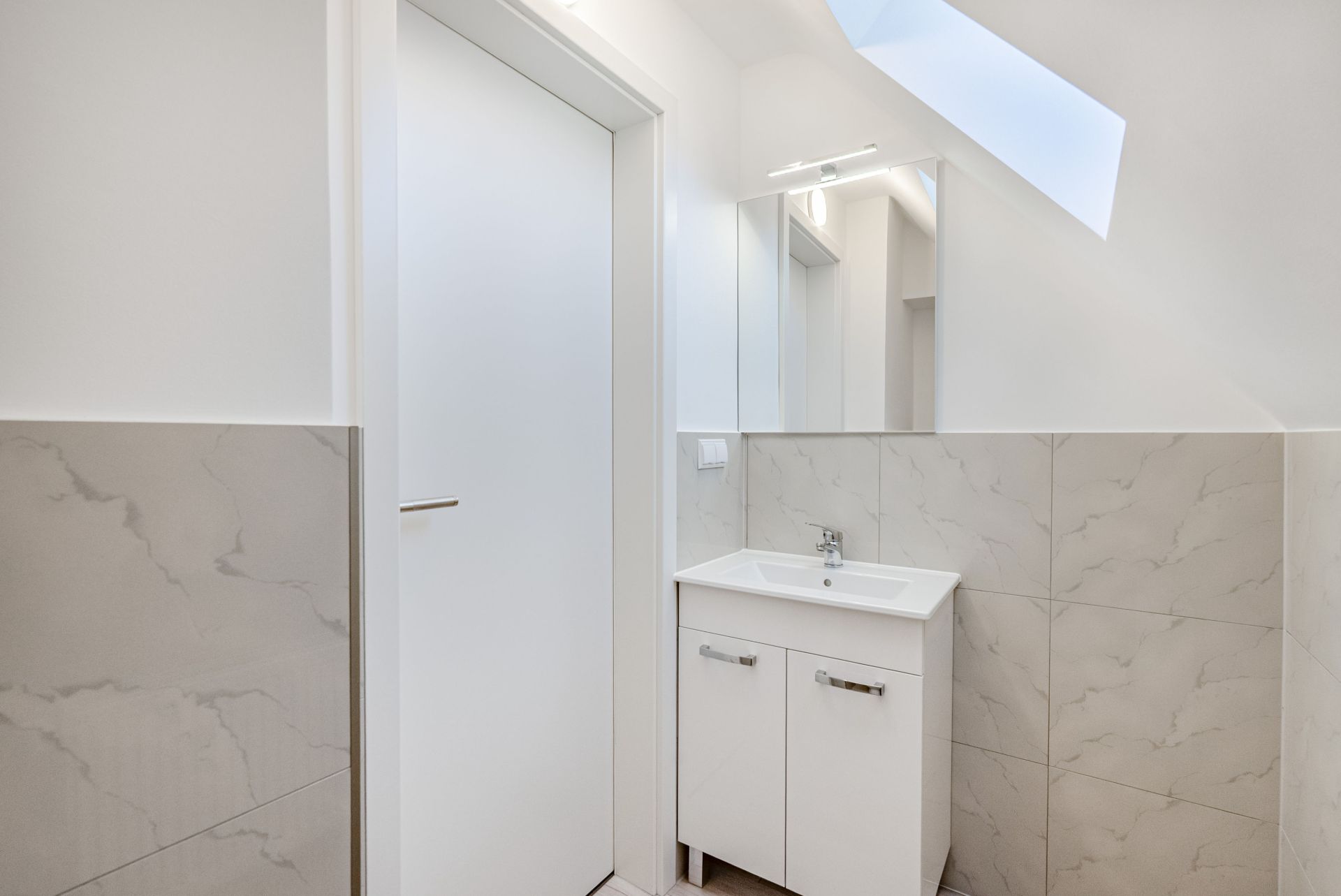 Modernes Badezimmer mit Waschbecken, Spiegel und weißer Tür unter Dachfenster.