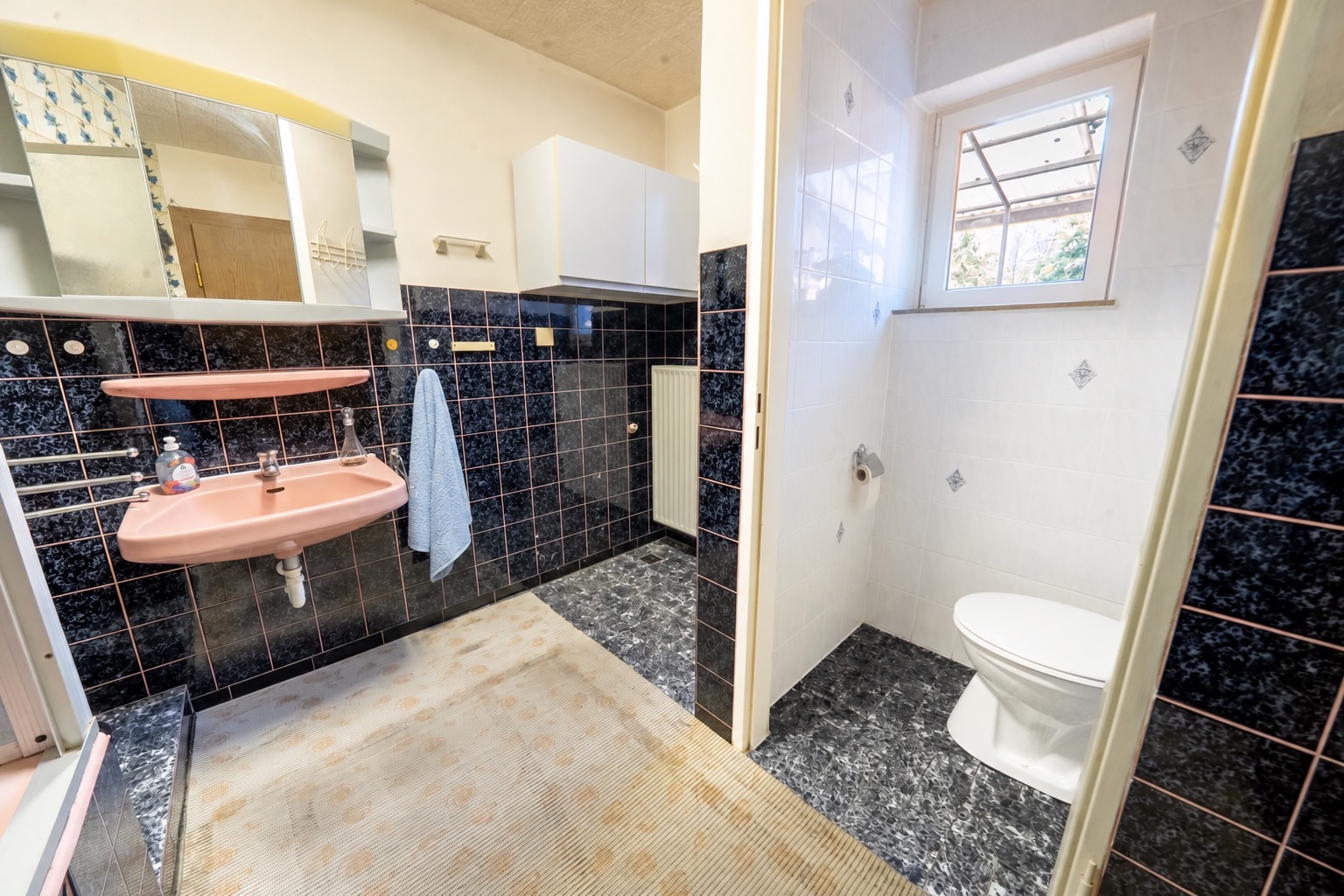 Badezimmer mit rosa Waschbecken, Spiegel, Handtuch und separatem WC-Bereich mit Fenster.