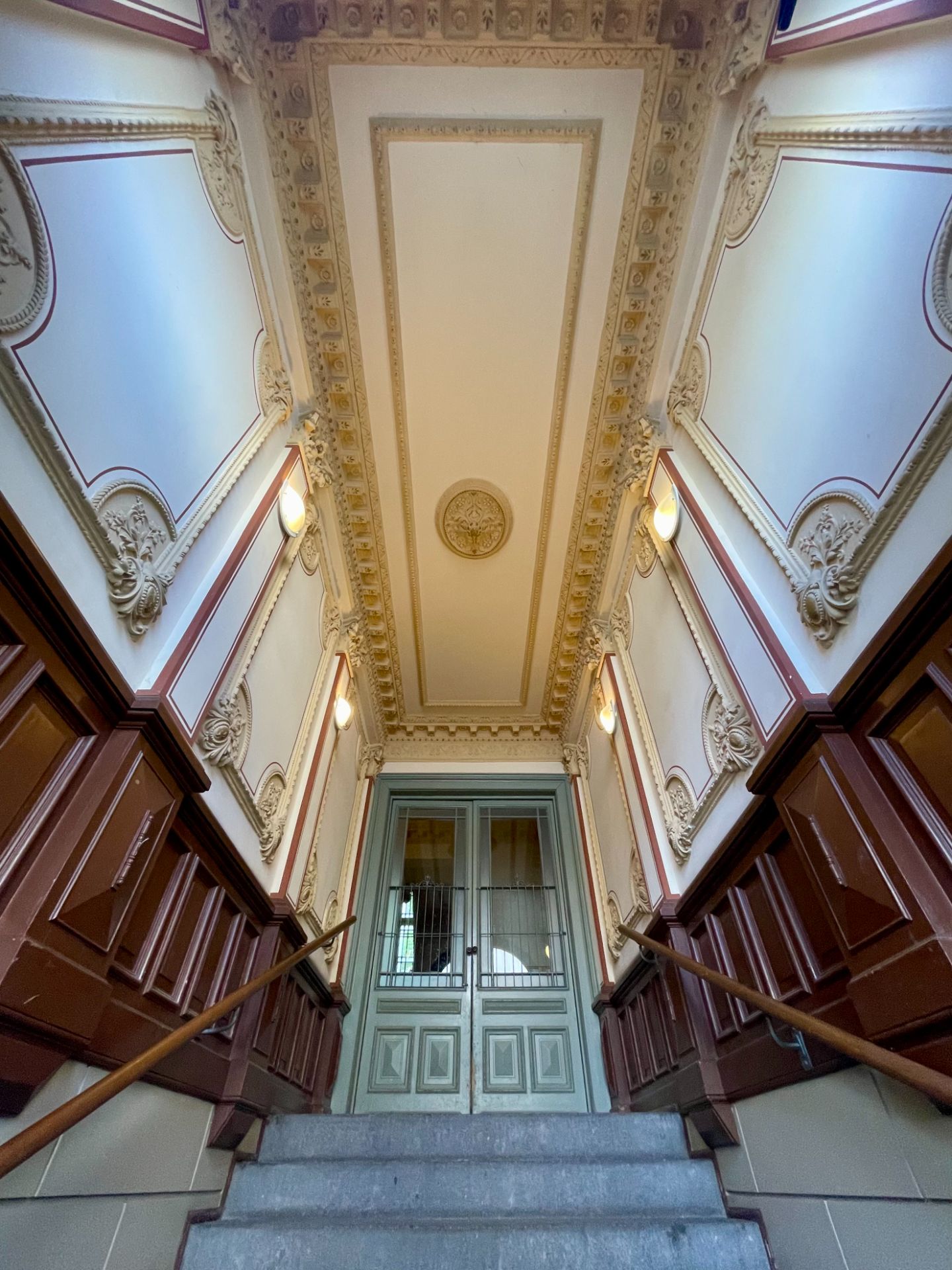 Verzierte Treppenhausdecke mit Wandverzierungen und Doppeltür am Ende der Treppe.