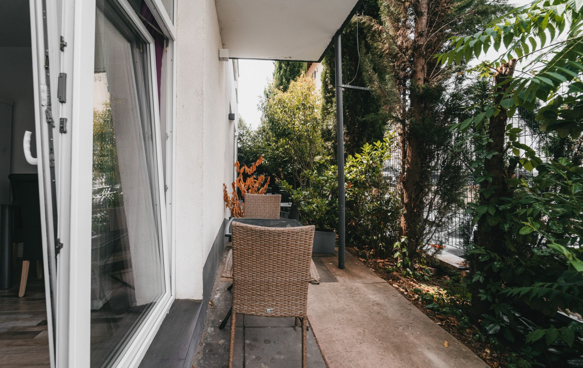 Kleiner Balkon mit zwei Stühlen und Tisch, umgeben von Pflanzen und Bäumen.