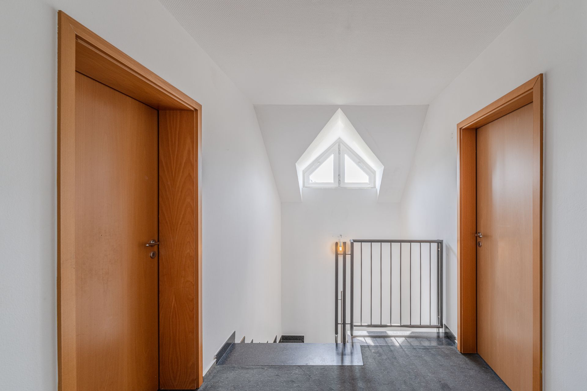 Flur mit zwei Holztüren, Treppengeländer und dreieckigem Fenster am Ende.