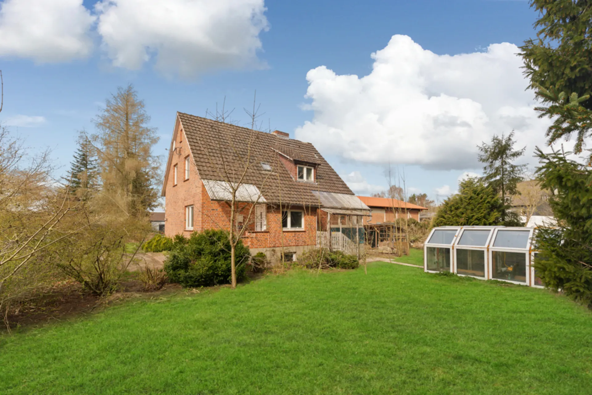 Rotes Backsteinhaus mit Satteldach und Wintergarten, umgeben von grünem Garten und Bäumen.
