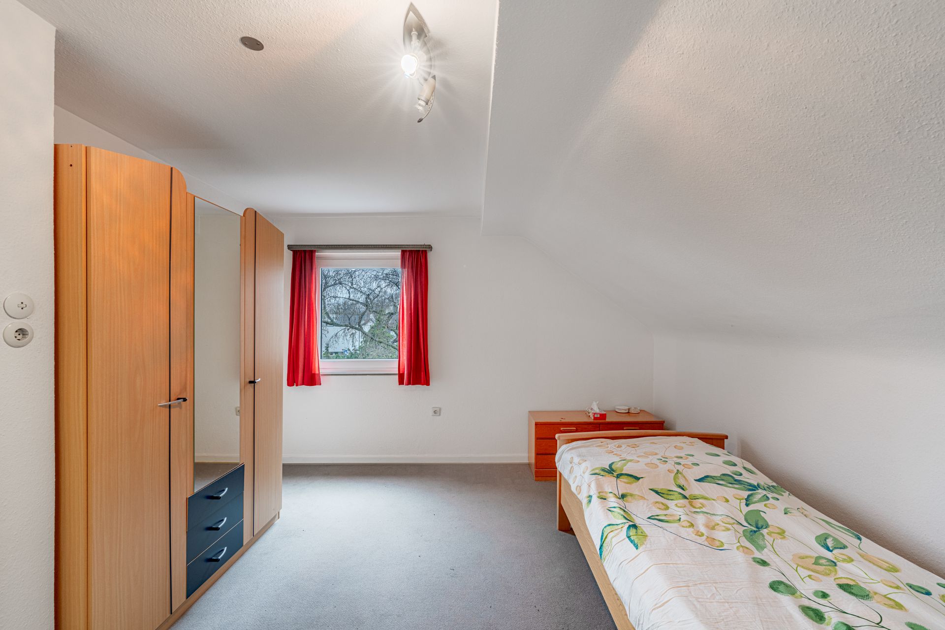 Schlichtes Schlafzimmer mit Einzelbett, Kleiderschrank, Kommode und Fenster mit roten Vorhängen.