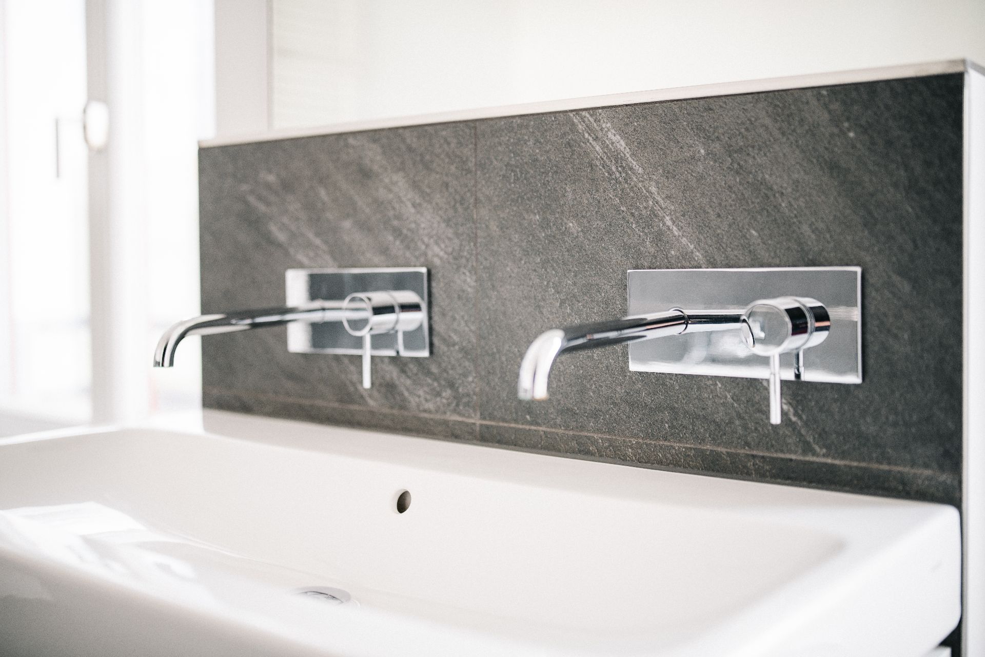 Zwei moderne Wandarmaturen über einem weißen Waschbecken mit dunkler Fliesenrückwand.