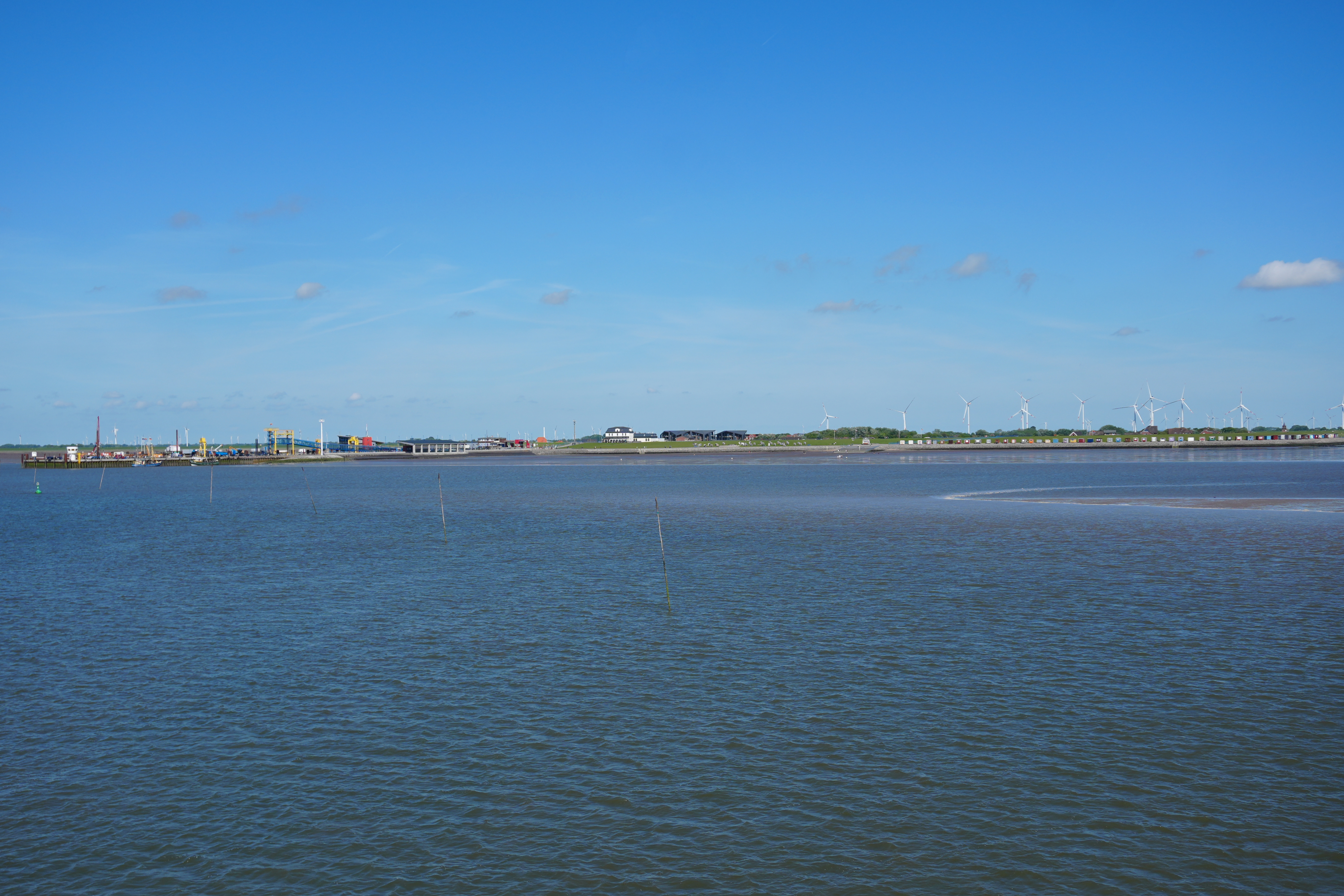 Blick auf ruhiges Wasser mit Windrädern und Gebäuden am fernen Ufer unter blauem Himmel.