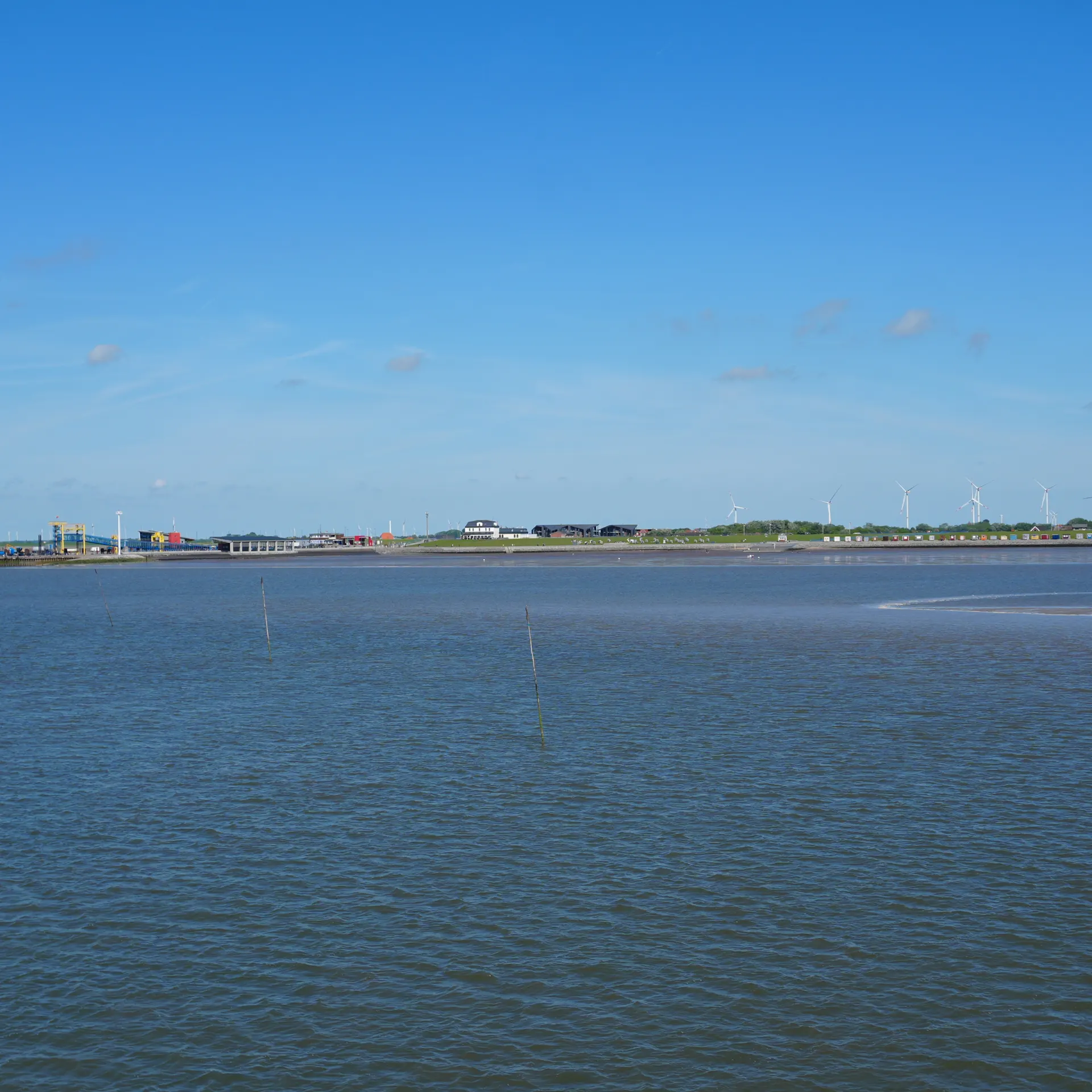 Blick auf ruhiges Wasser mit Windrädern und Gebäuden am fernen Ufer unter blauem Himmel.