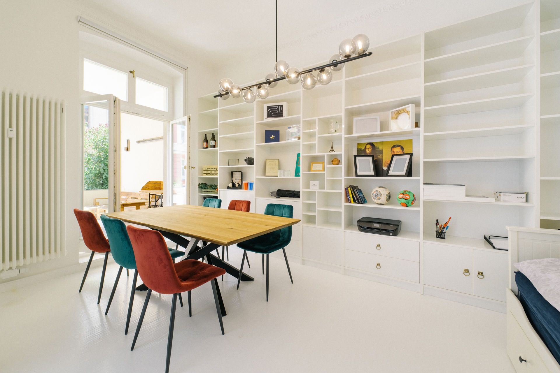 Helles Zimmer mit Holztisch, bunten Stühlen und weißem Regal an der Wand.