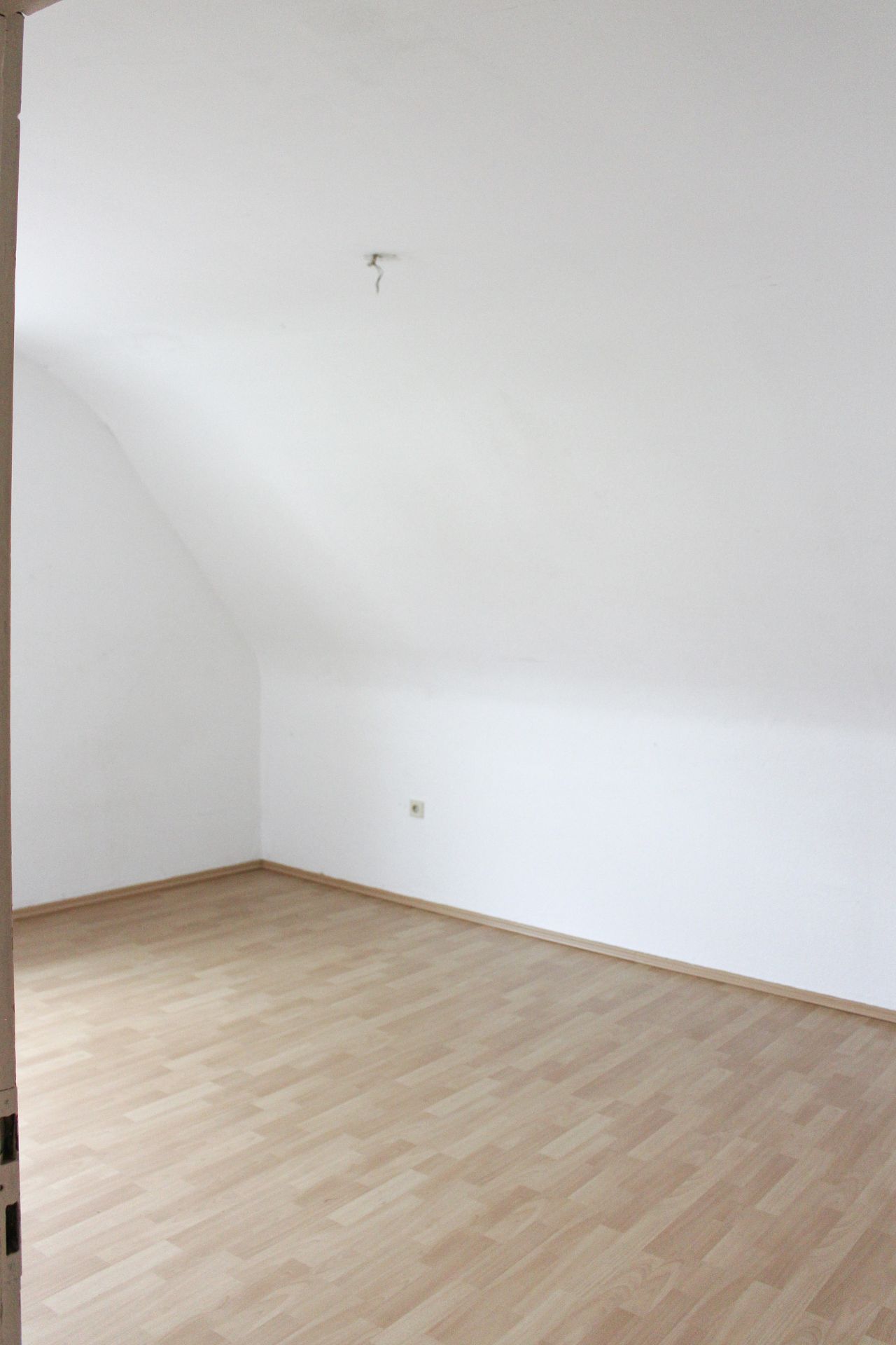 Leerer Raum mit hellen Wänden und schräger Decke, heller Laminatboden.