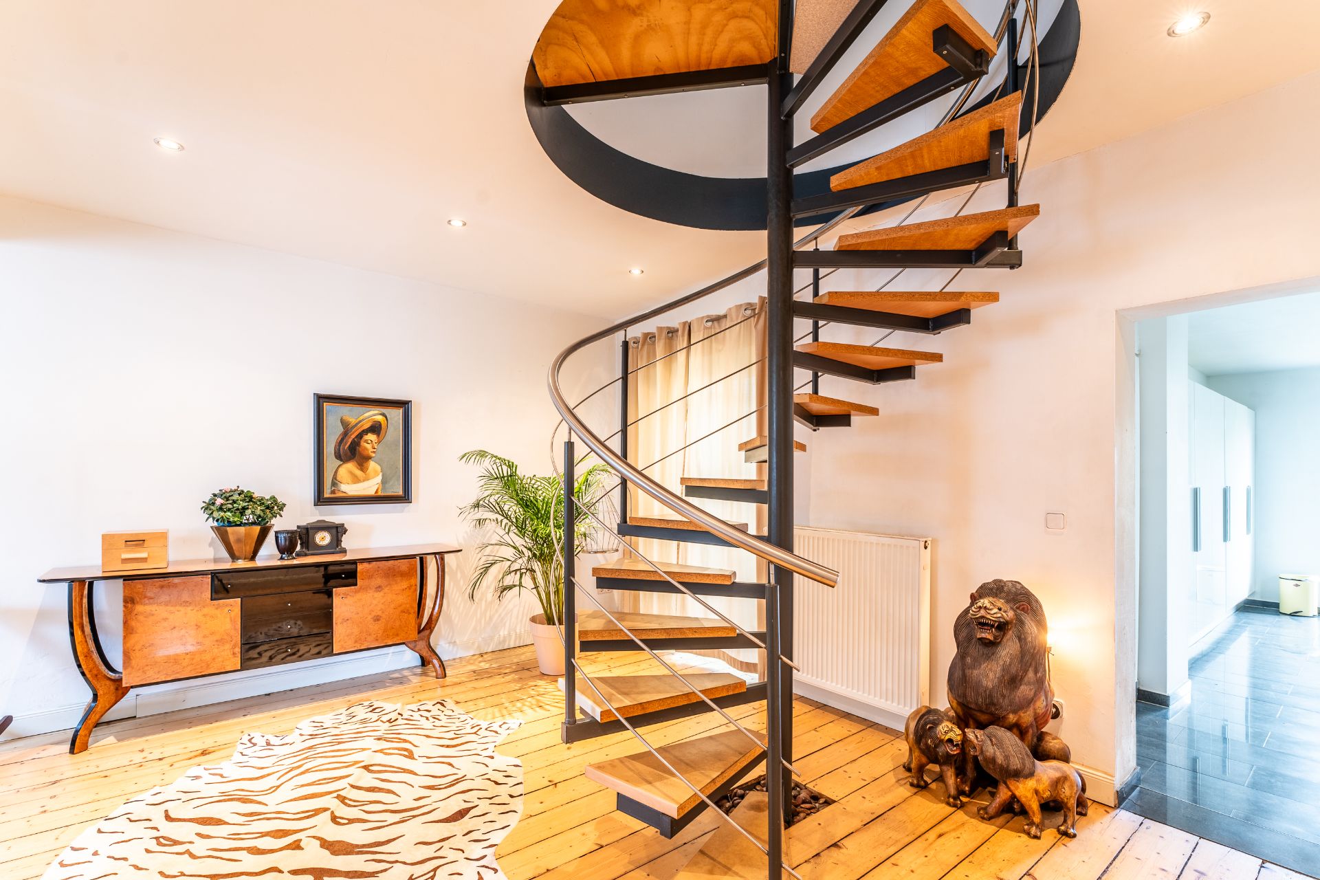 Wohnzimmer mit Wendeltreppe, Holzfußboden, Kommode, Gemälde und Löwenfiguren.