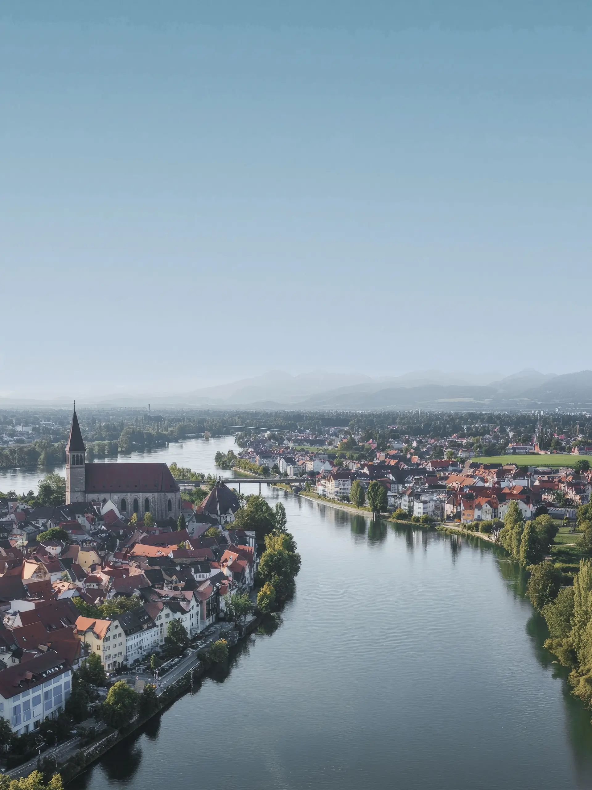 Luftaufnahme einer Stadt mit Fluss, Kirche und roten Dächern bei klarem Himmel.