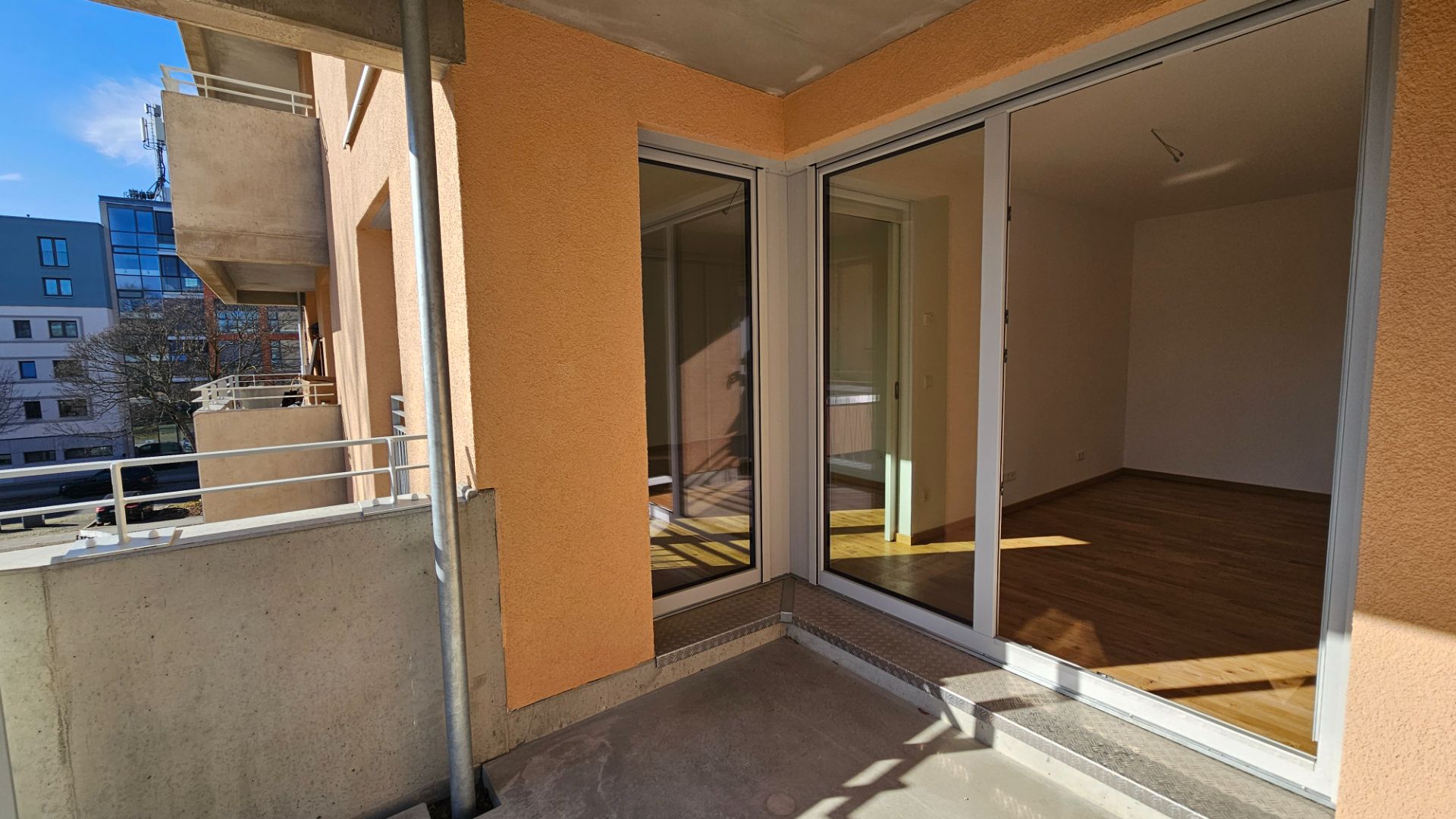 Balkon mit Glasgeländer und Zugang zu einem Zimmer mit Holzfußboden in einem Wohngebäude.