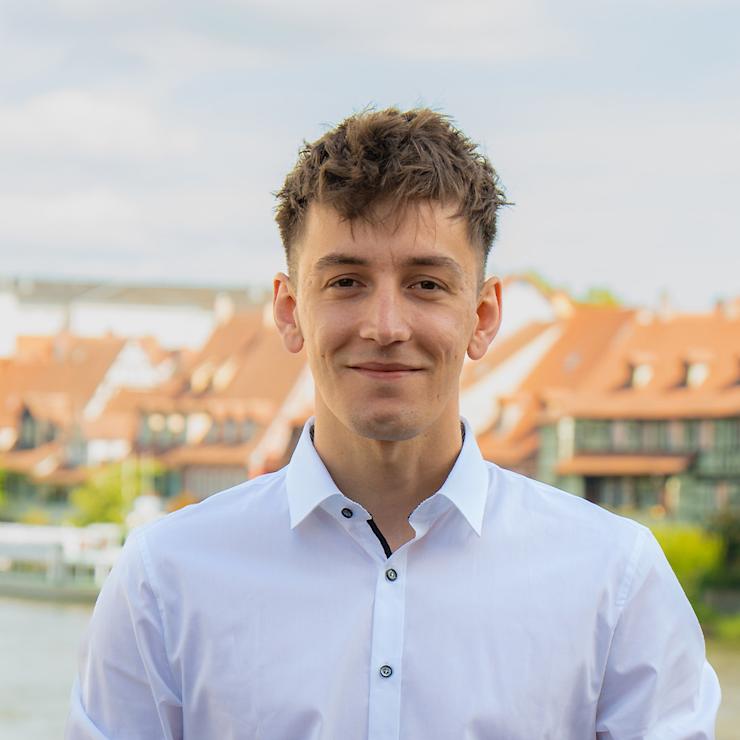 Ein Mann mit weißem Hemd steht draußen vor Gebäuden mit roten Dächern.