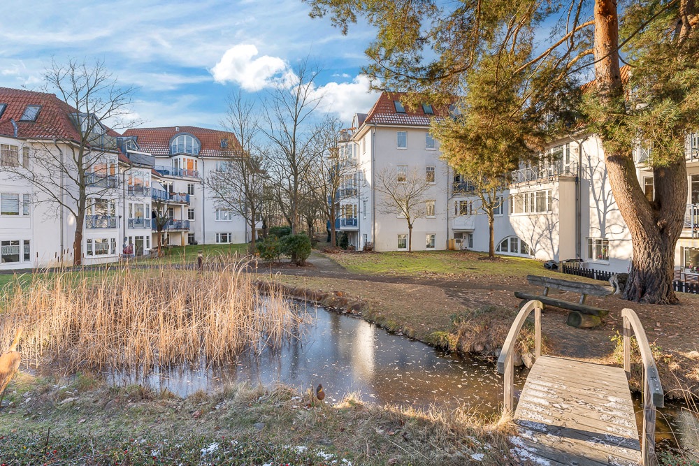 Wohnhäuser mit roten Dächern, Gartenteich, Holzbrücke und Bäumen im Innenhof.
