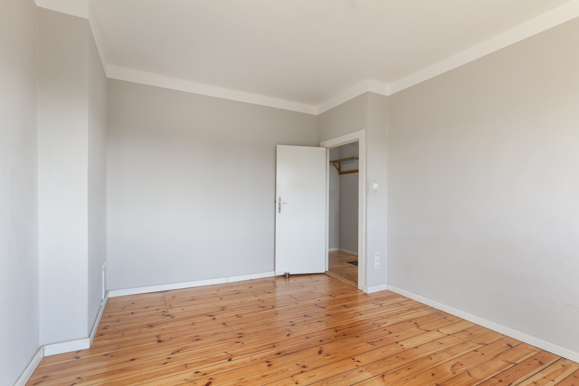 Leeres Zimmer mit Holzboden, grauen Wänden und geöffneter Tür zu einem kleinen Nebenraum.