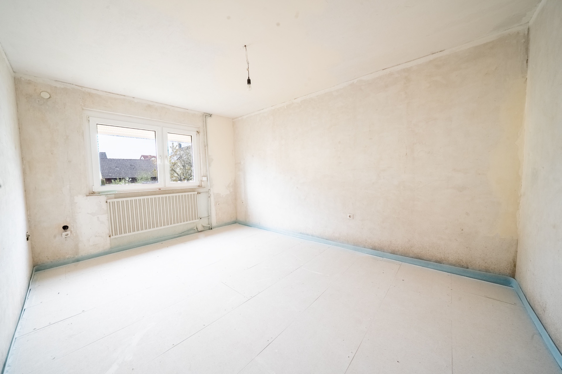 Leeres, renovierungsbedürftiges Zimmer mit zwei Fenstern und einem Heizkörper an der Wand.