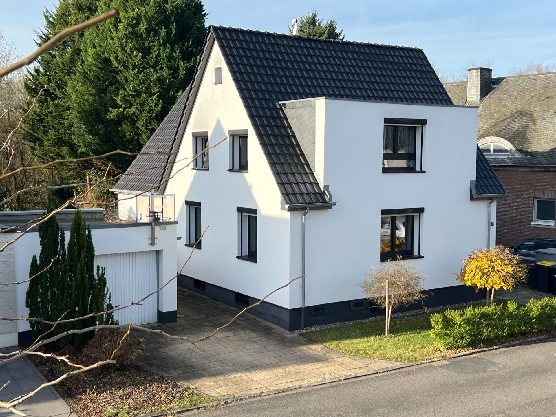 Weißes Einfamilienhaus mit dunklem Dach, Garage und kleinem Garten an einer Straße.