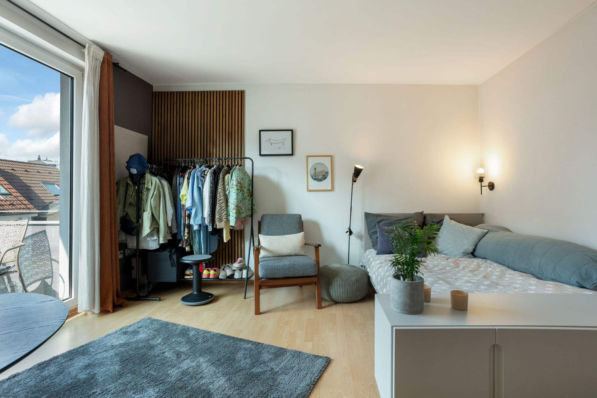 Helles Schlafzimmer mit Bett, Sessel, Teppich, Kleiderstange und Balkonzugang.