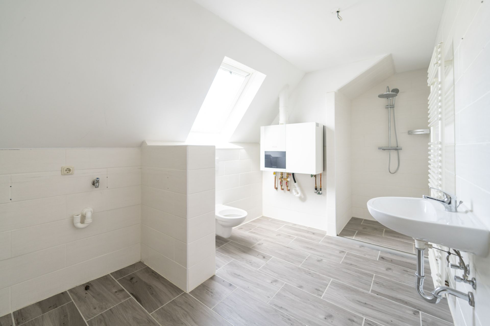 Helles Badezimmer mit Dachfenster, WC, Waschbecken und offener Dusche auf Holzoptik-Fliesen.