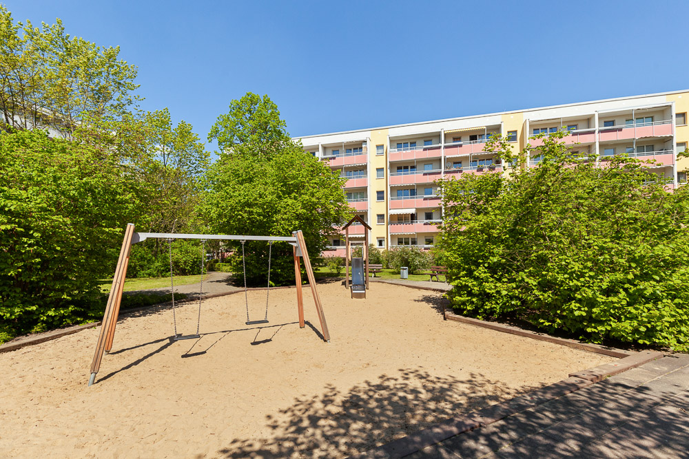 Spielplatz mit zwei Schaukeln im Sand vor einem Wohnhaus und Bäumen.
