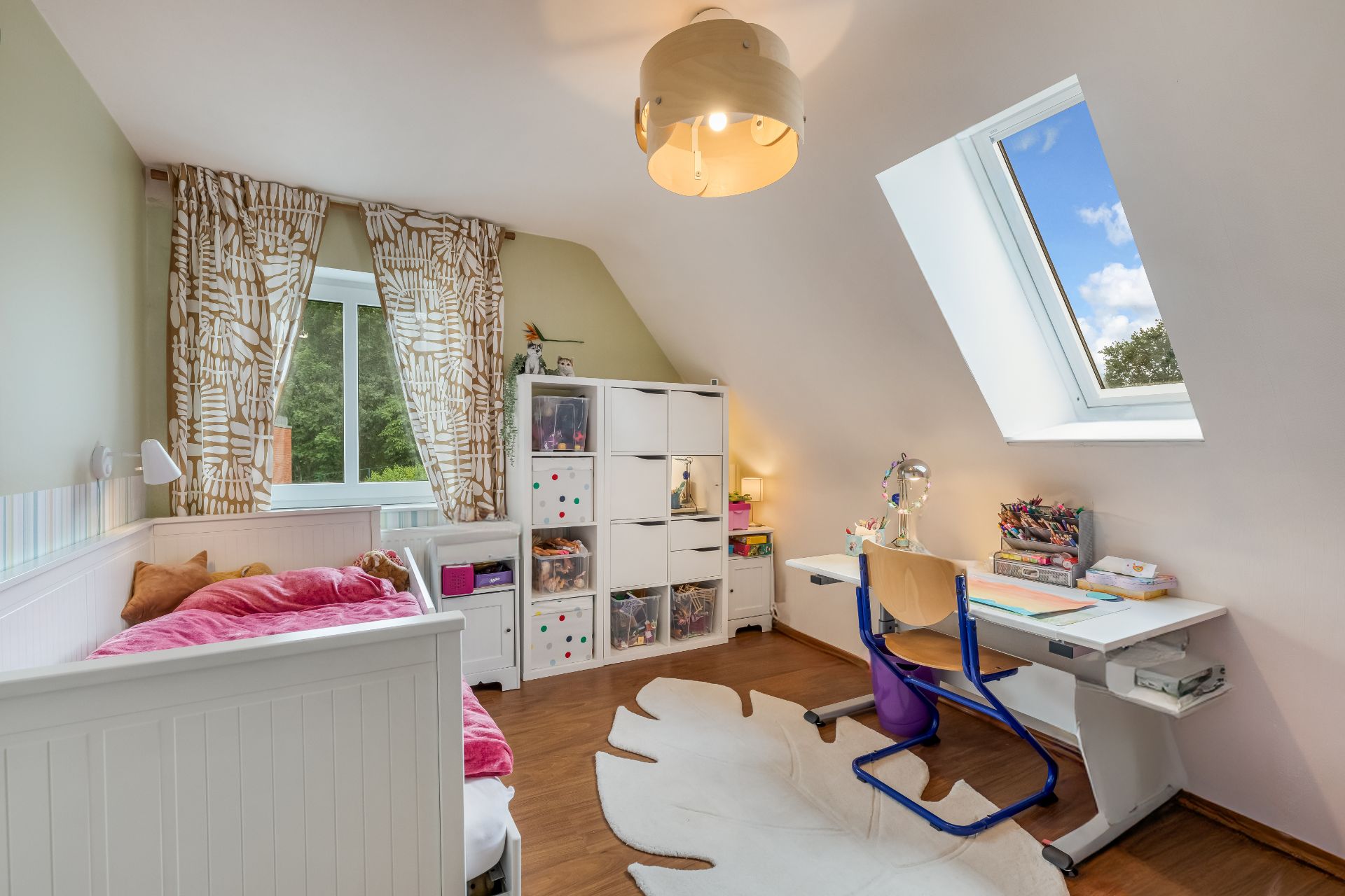 Helles Kinderzimmer mit Bett, Schreibtisch, Regal und zwei Fenstern.
