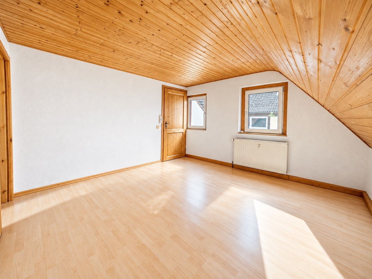Heller, leerer Raum mit Holzdecke, zwei Fenstern, Tür und Heizung.