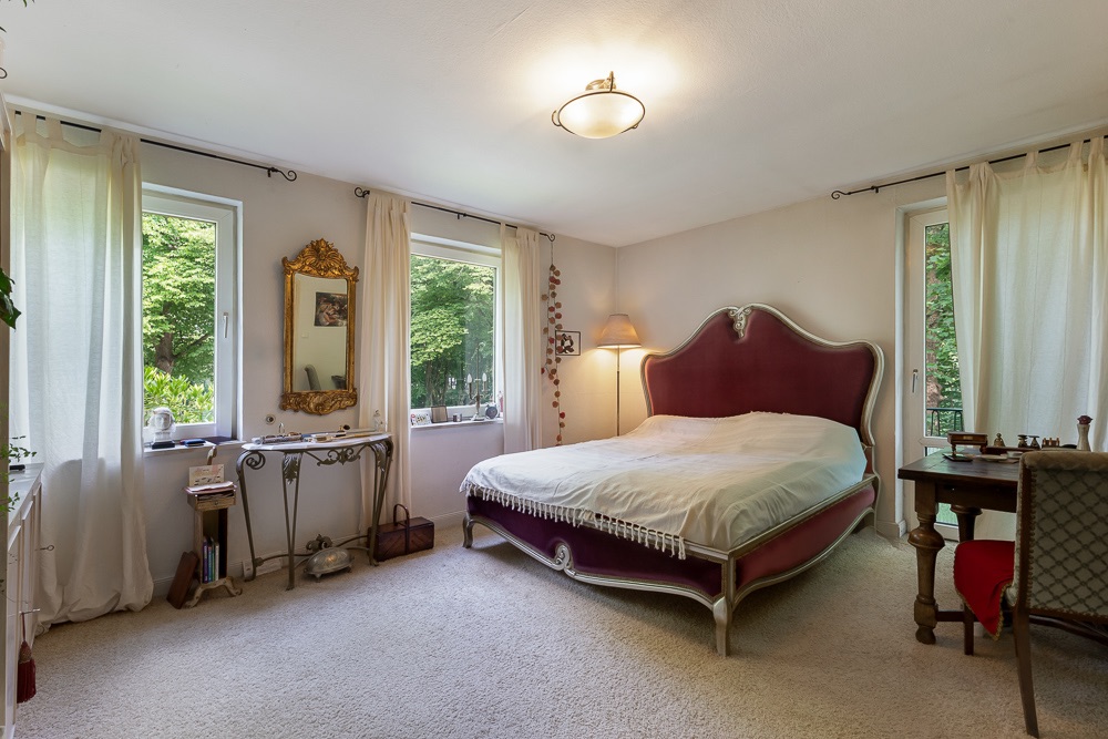 Helles Schlafzimmer mit rotem Bett, Schreibtisch, Spiegel und großen Fenstern mit Vorhängen.