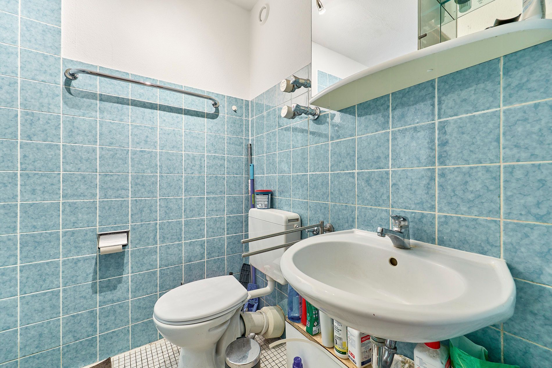 Kleines Badezimmer mit blauen Fliesen, Waschbecken, Toilette und Ablage mit Putzmitteln.