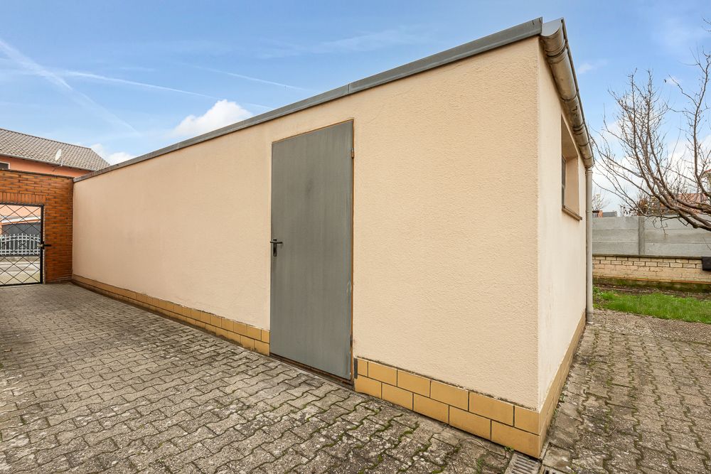 Beiger Schuppen mit grauer Tür auf gepflastertem Hof, daneben etwas Rasen.