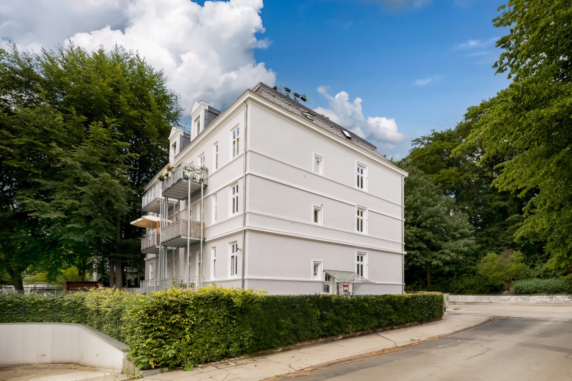 Weißes Mehrfamilienhaus mit Balkonen, umgeben von Bäumen und Hecken an einer ruhigen Straße.