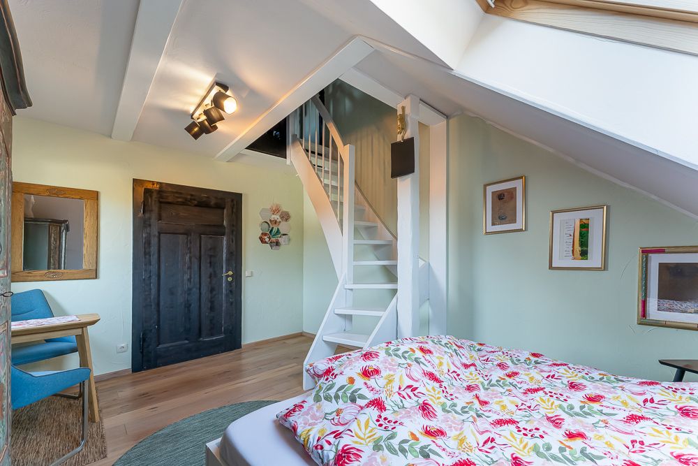Helles Schlafzimmer mit Blumenbettdecke, Schreibtisch, Holztreppe und Dachfenster.