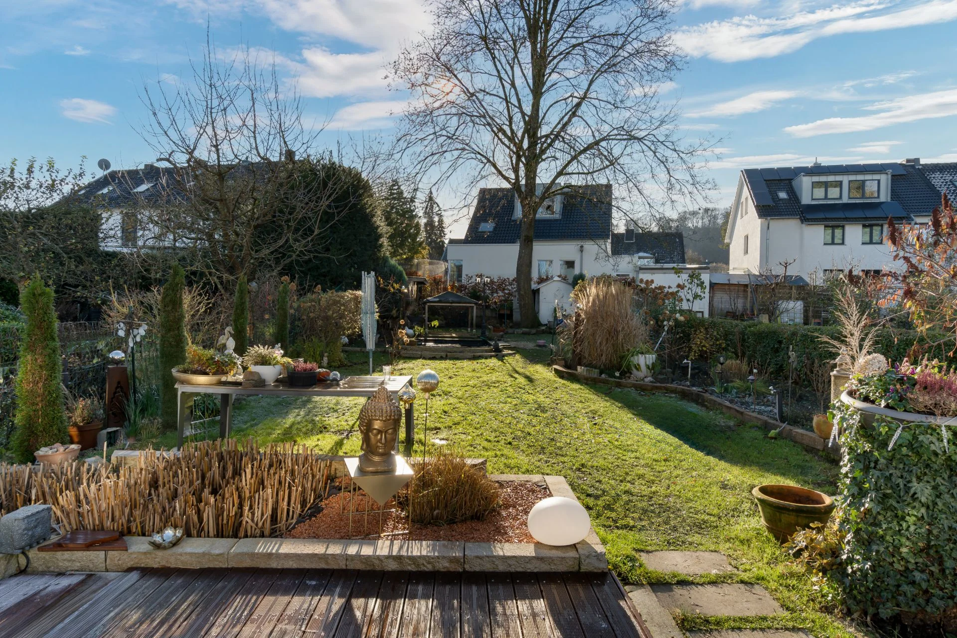 Garten mit Rasenfläche, Terrasse, Pflanzgefäßen und Bäumen neben Wohnhäusern bei sonnigem Wetter.
