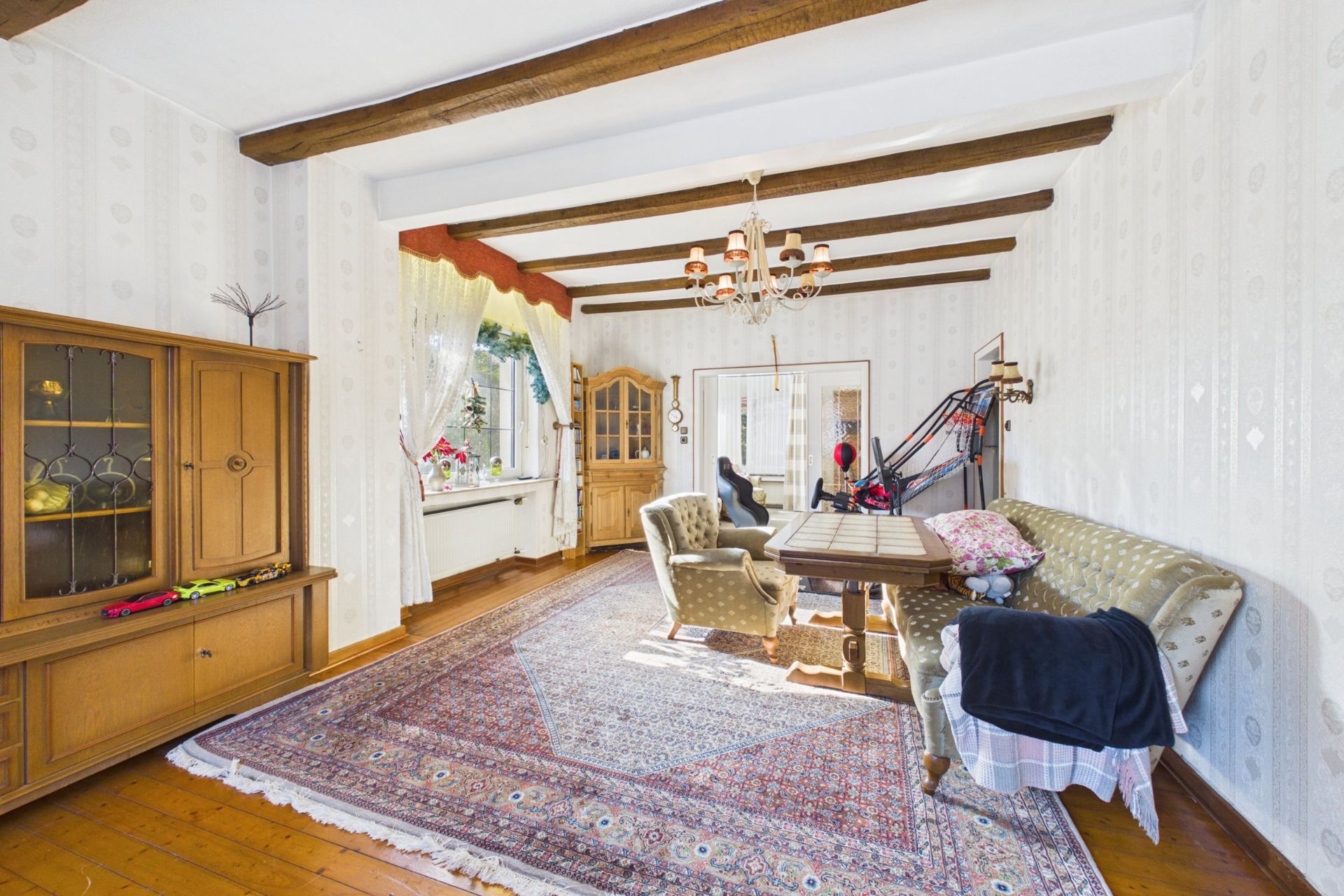 Helles Wohnzimmer mit Teppich, Holzbalken, Sofa, Tisch, Schrank und Fenster mit Vorhängen.