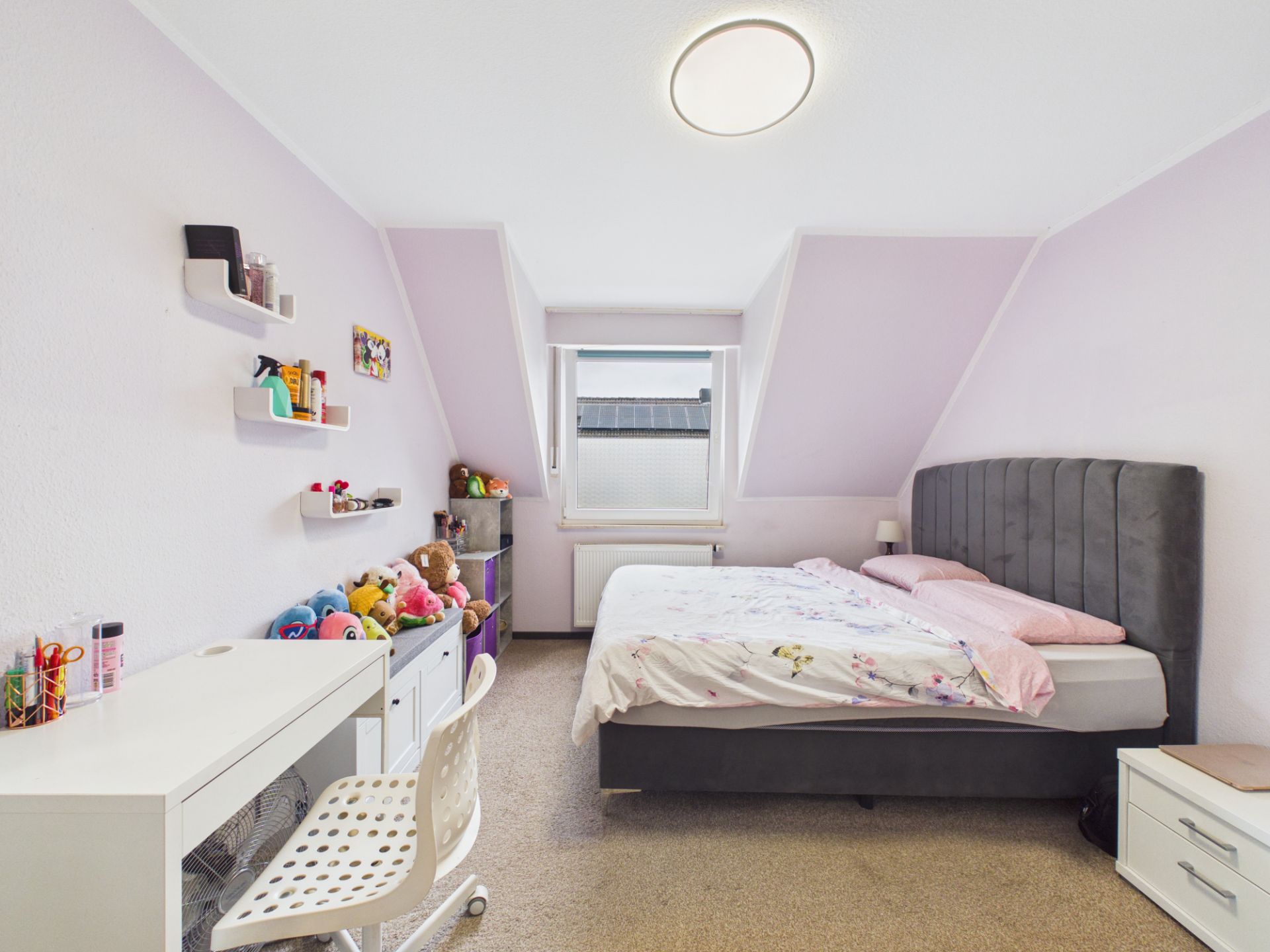 Helles Schlafzimmer mit Doppelbett, Schreibtisch, Stuhl, Regalen und Plüschtieren vor einem Fenster.