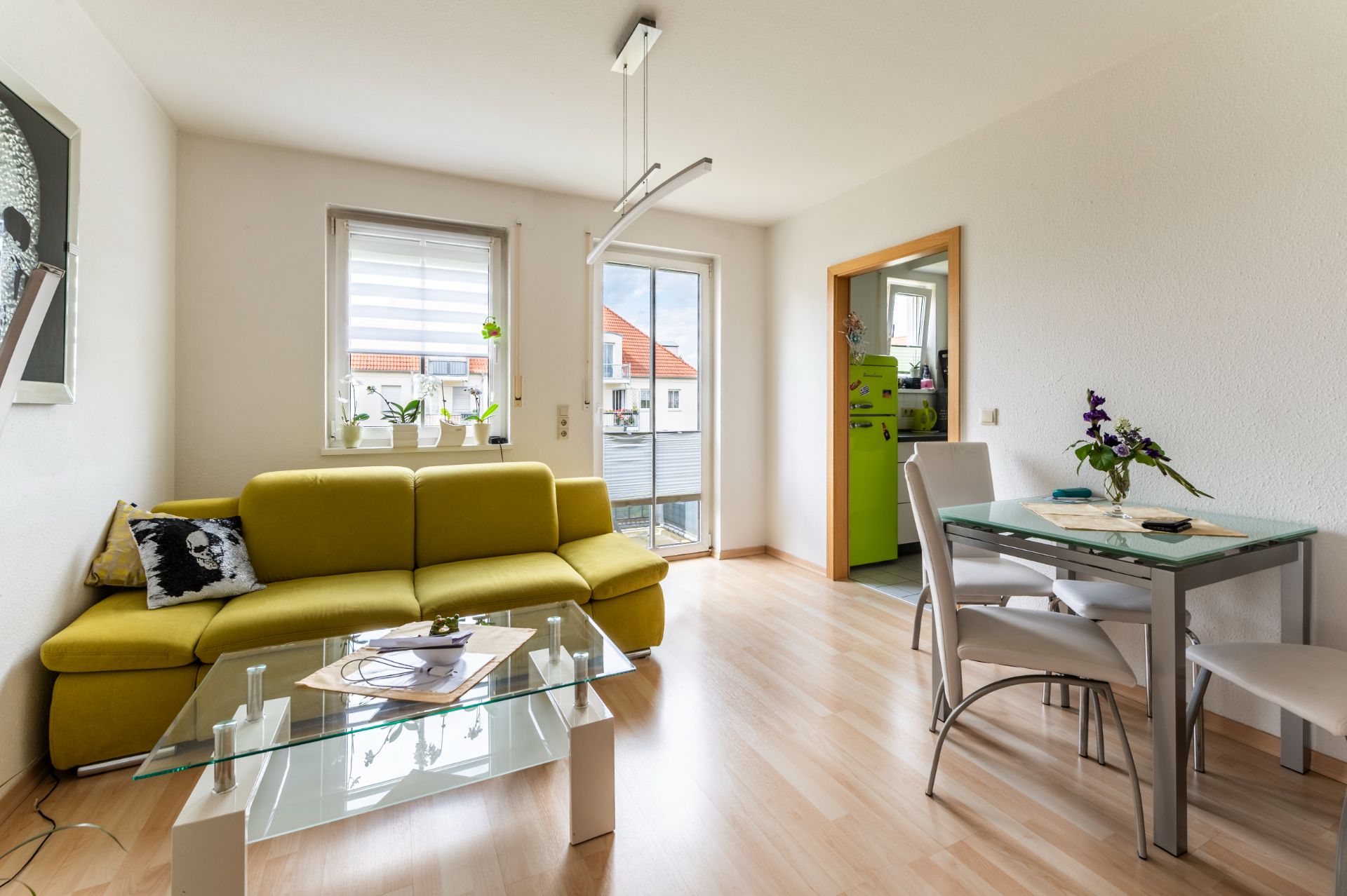Wohnzimmer mit gelbem Sofa, Glastisch, Esstisch mit vier Stühlen und Zugang zum Balkon.