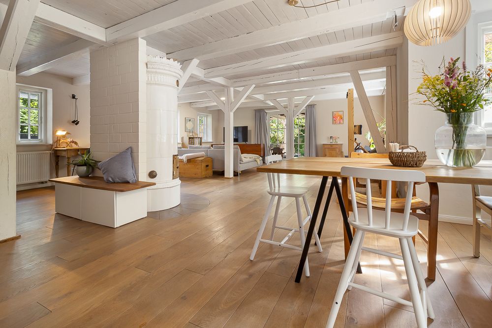 Helles Wohnzimmer mit Holzboden, Esstisch, weißen Balken und großem Kachelofen.
