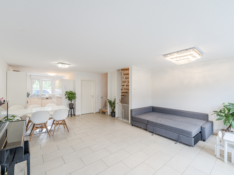 Helles Wohnzimmer mit grauem Sofa, Esstisch, weißen Stühlen und Pflanzen, offene Küche im Hintergrund.