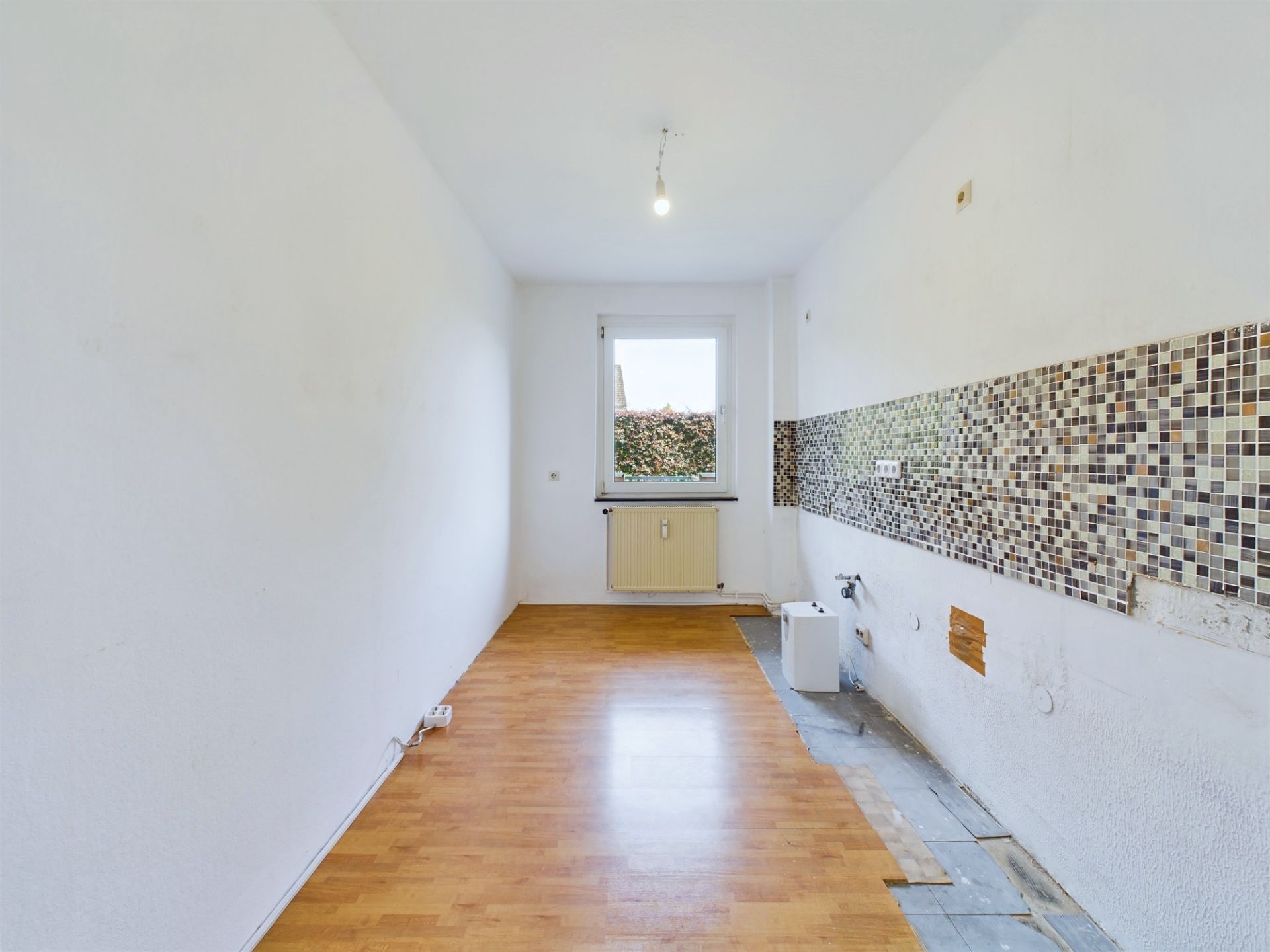 Leerer Raum mit Laminatboden, Fliesenspiegel an einer Wand und Fenster am Ende.