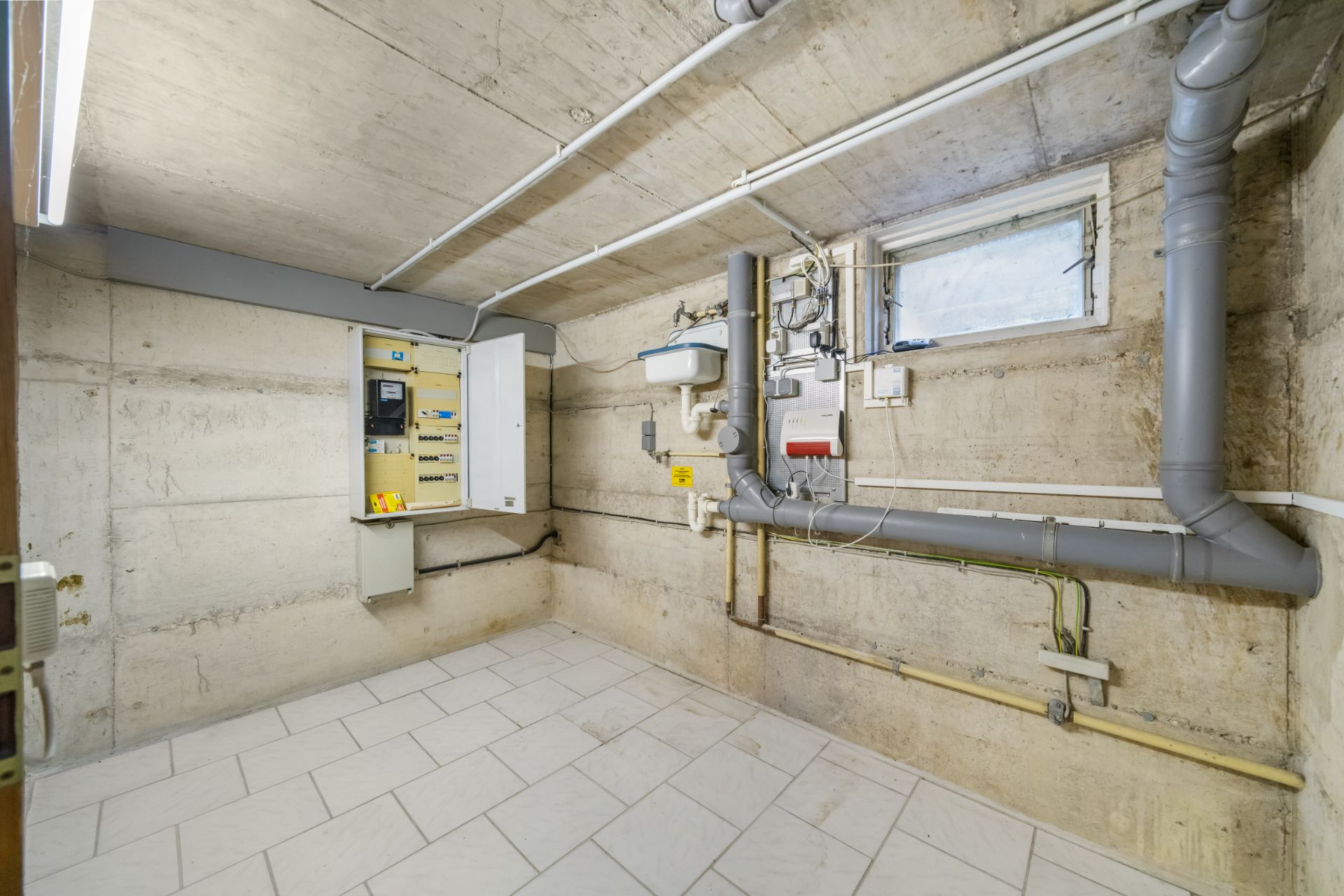 Kellerraum mit Fliesenboden, offenem Schaltschrank, Rohren und kleinem Fenster an der Wand.