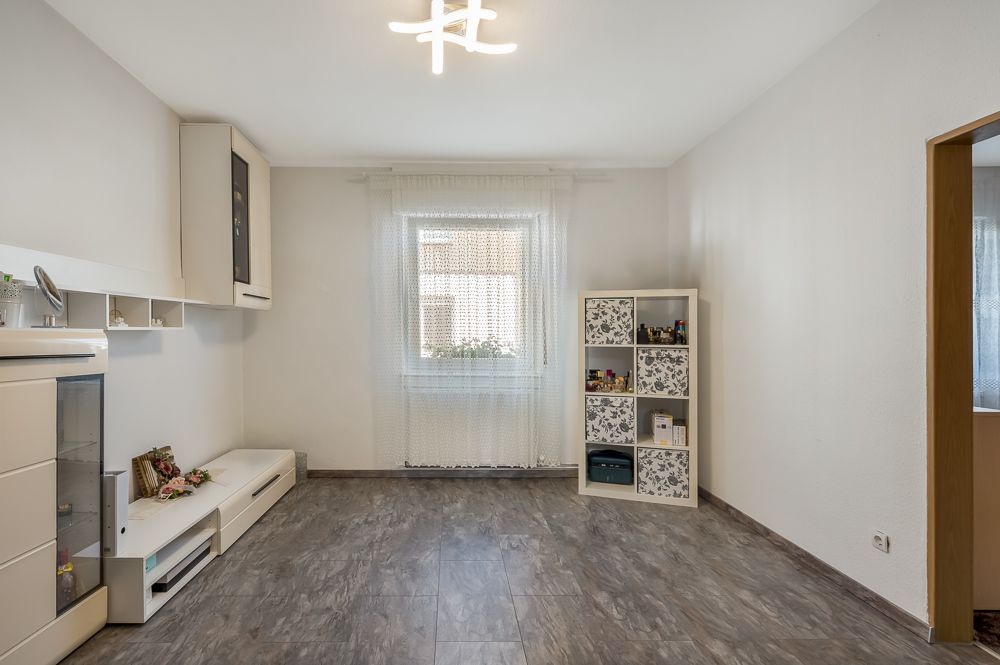 Helles Wohnzimmer mit Regal, Sideboard, Fenster mit Vorhang und grauem Boden.