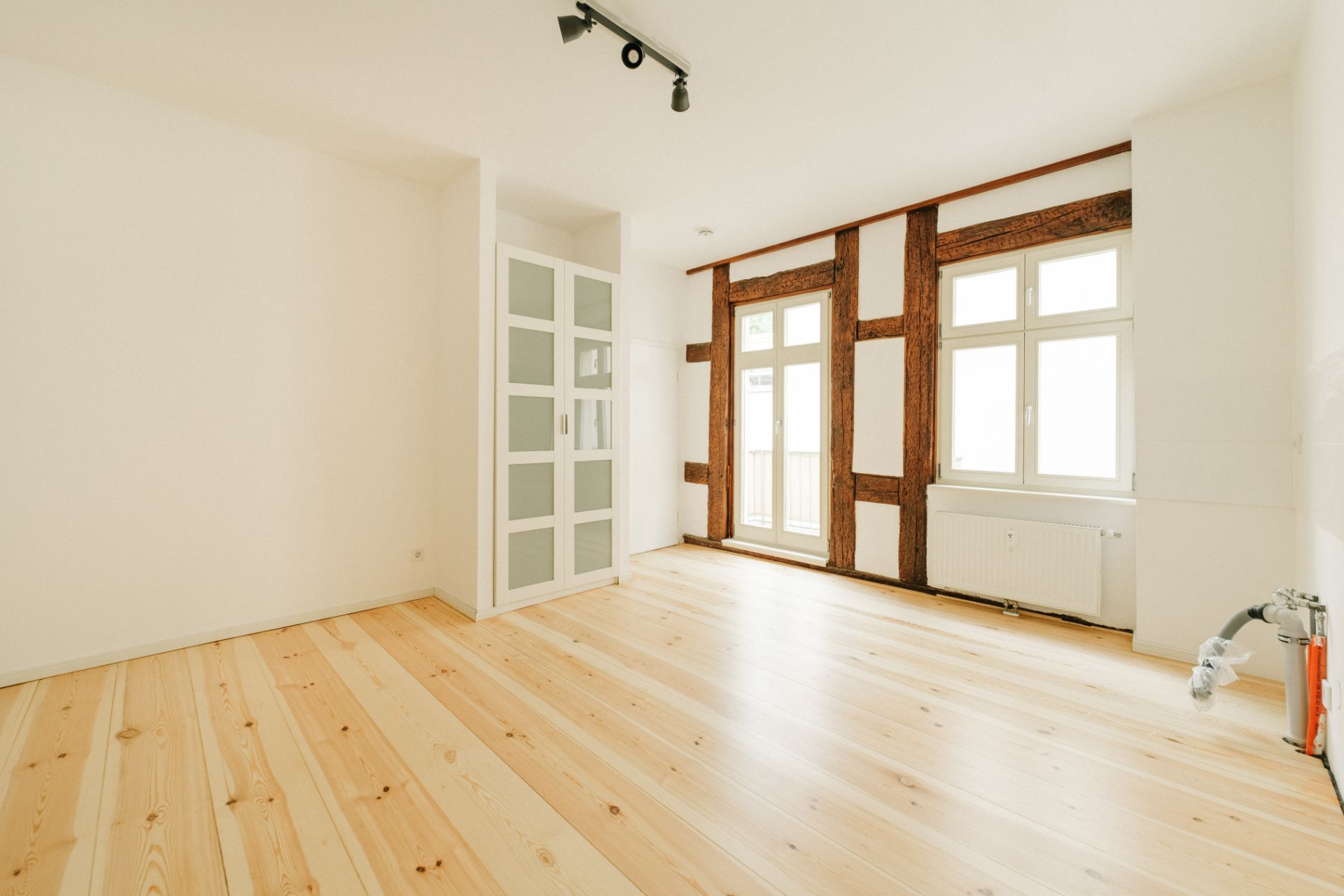 Heller Raum mit Holzboden, Fachwerkwand, großen Fenstern und einer weißen Tür mit Glaseinsatz.