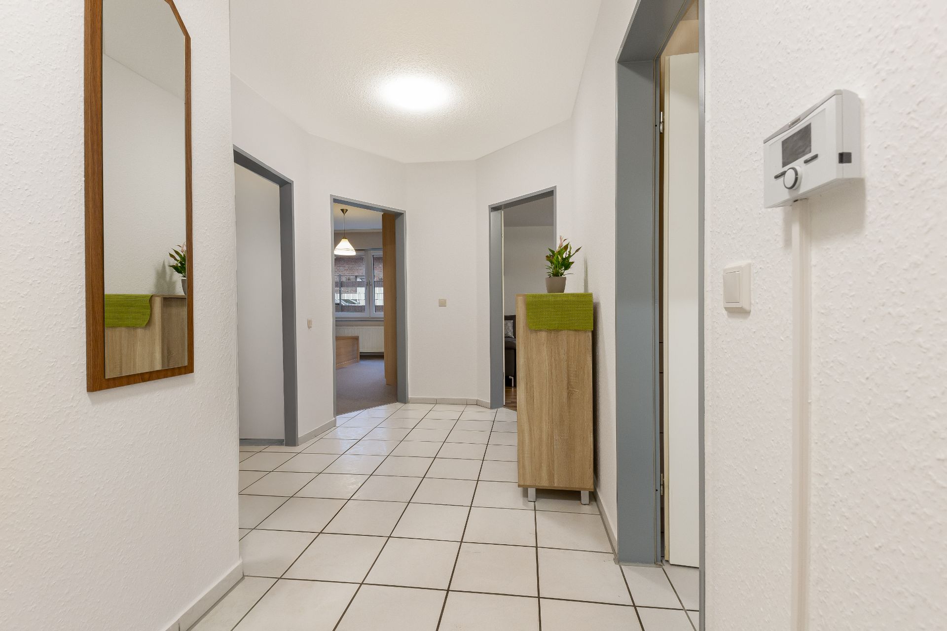 Heller Flur mit weißen Fliesen, Spiegel, Kommode und mehreren geöffneten Türen zu anderen Räumen.