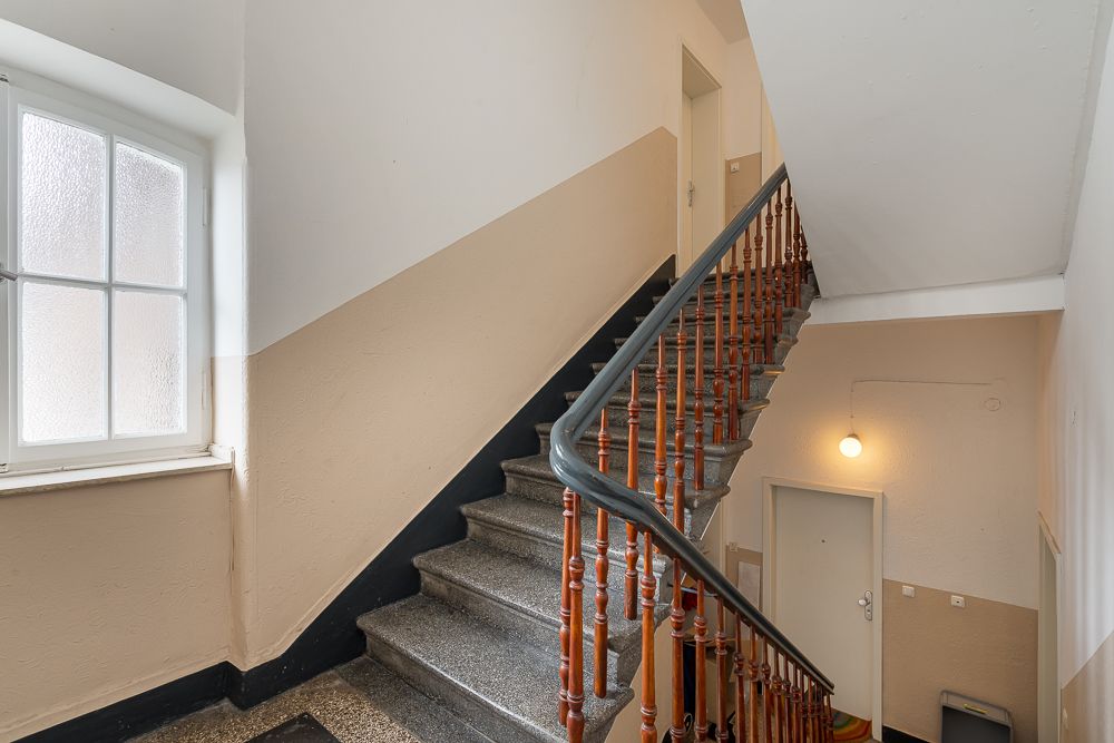 Treppenhaus mit Holzgeländer, Fenster und zwei Wohnungstüren unter künstlichem Licht.