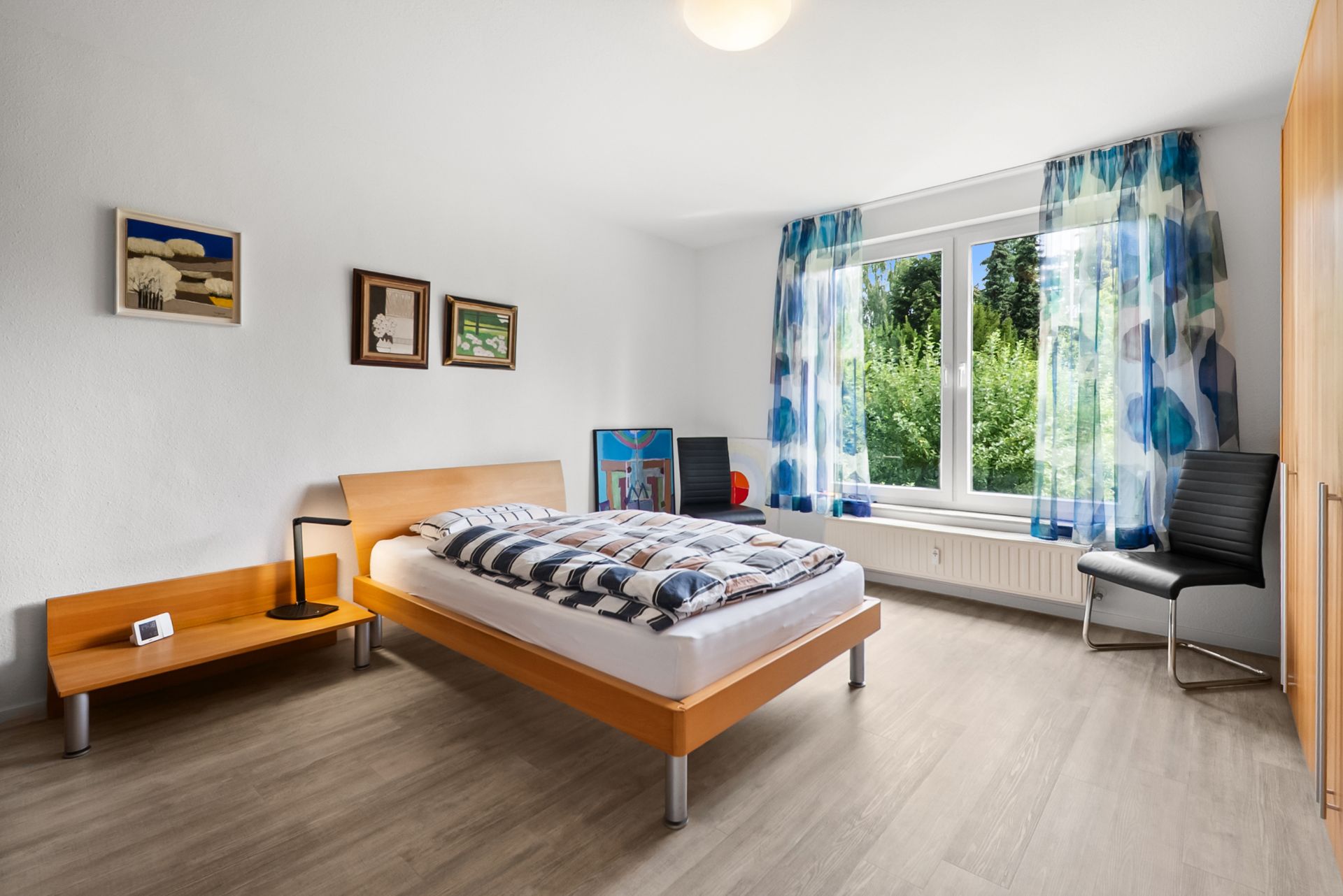 Helles Schlafzimmer mit Holzbett, Nachttisch, Stühlen, großem Fenster und bunten Vorhängen.